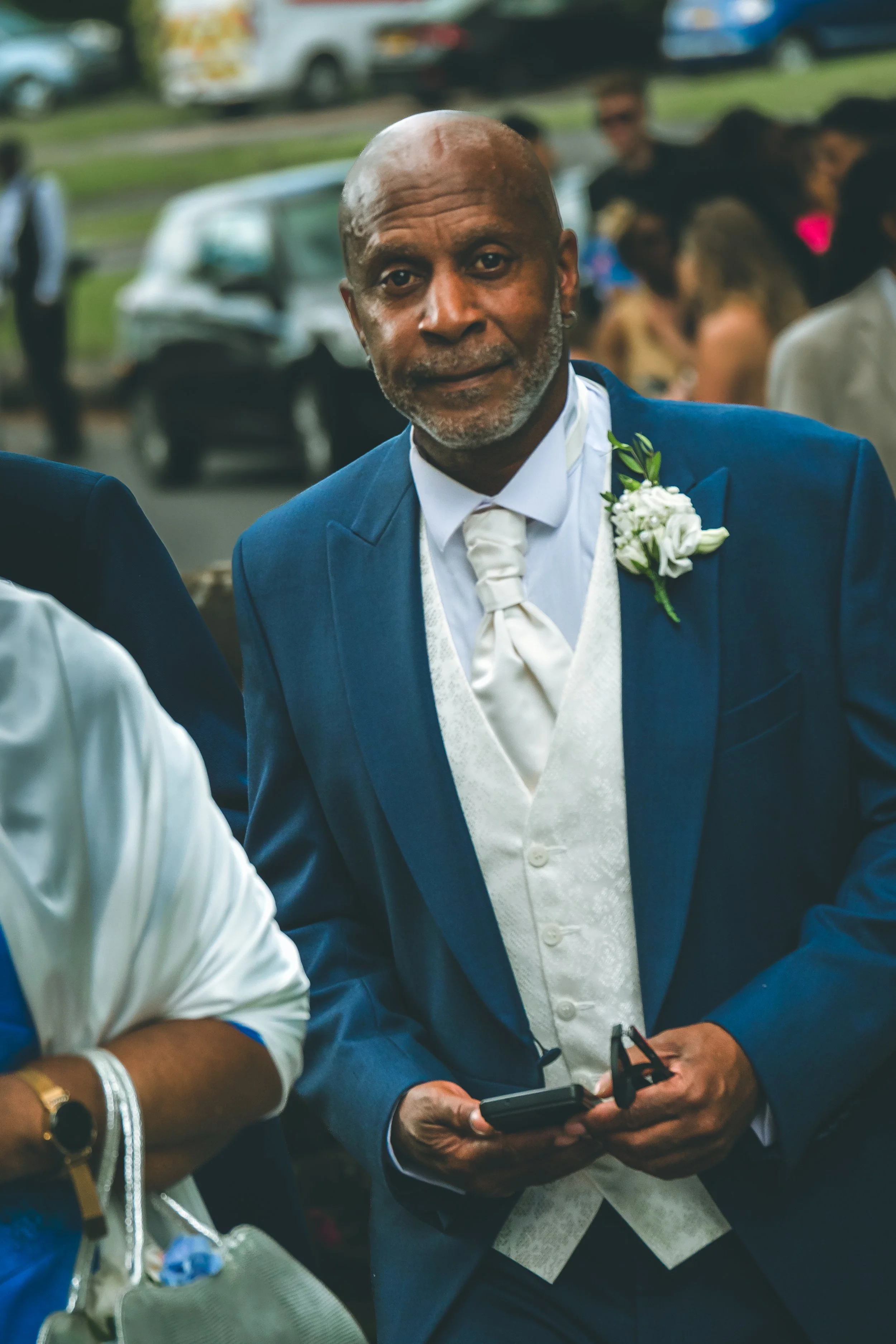 Bald man in blue suit wedding attire, holding a phone, standing outdoors with blurred people and cars in background.