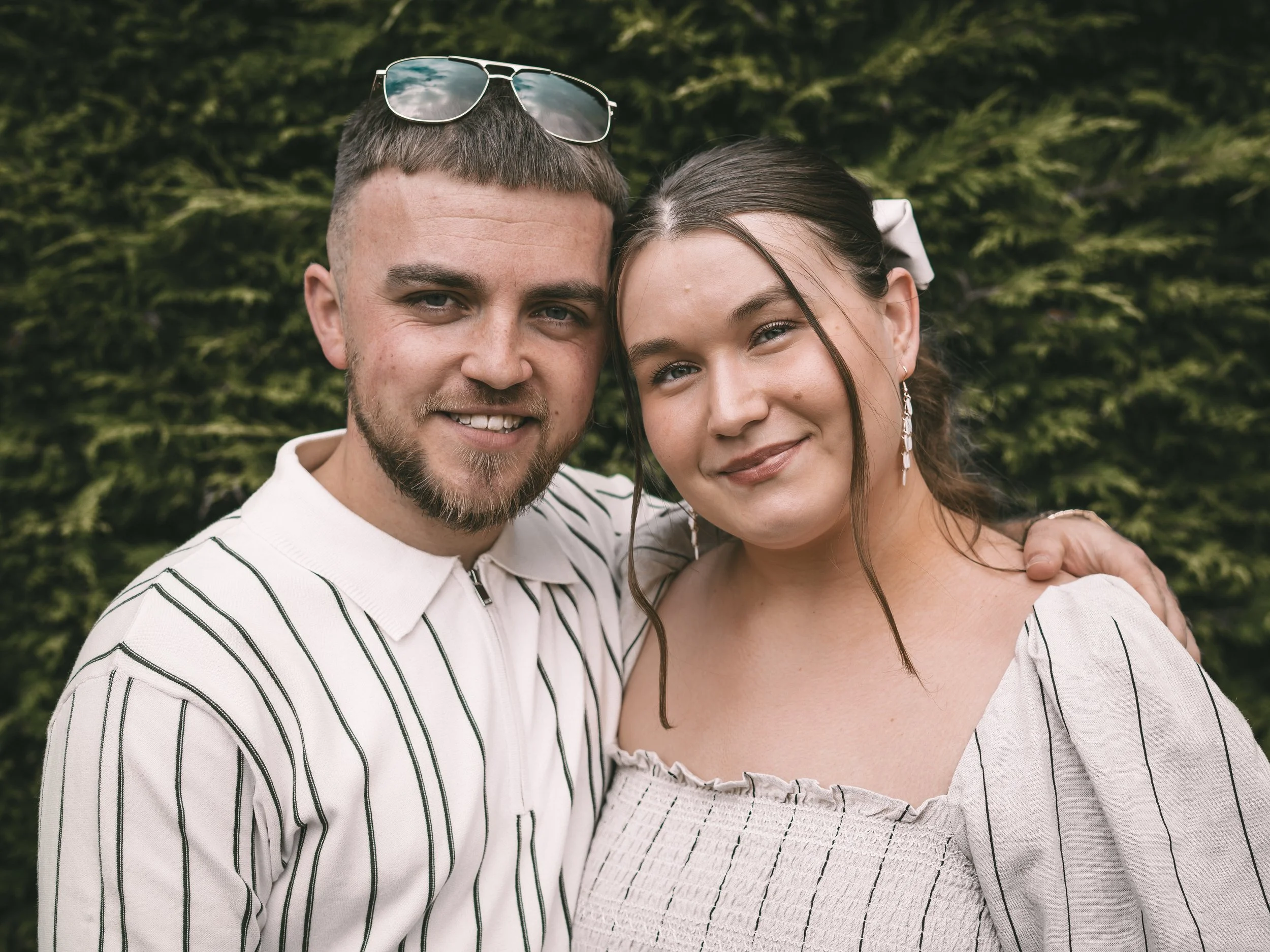 A young couple taking a selfie outdoors, with greenery in the background, smiling and close together, wearing light-colored tops.