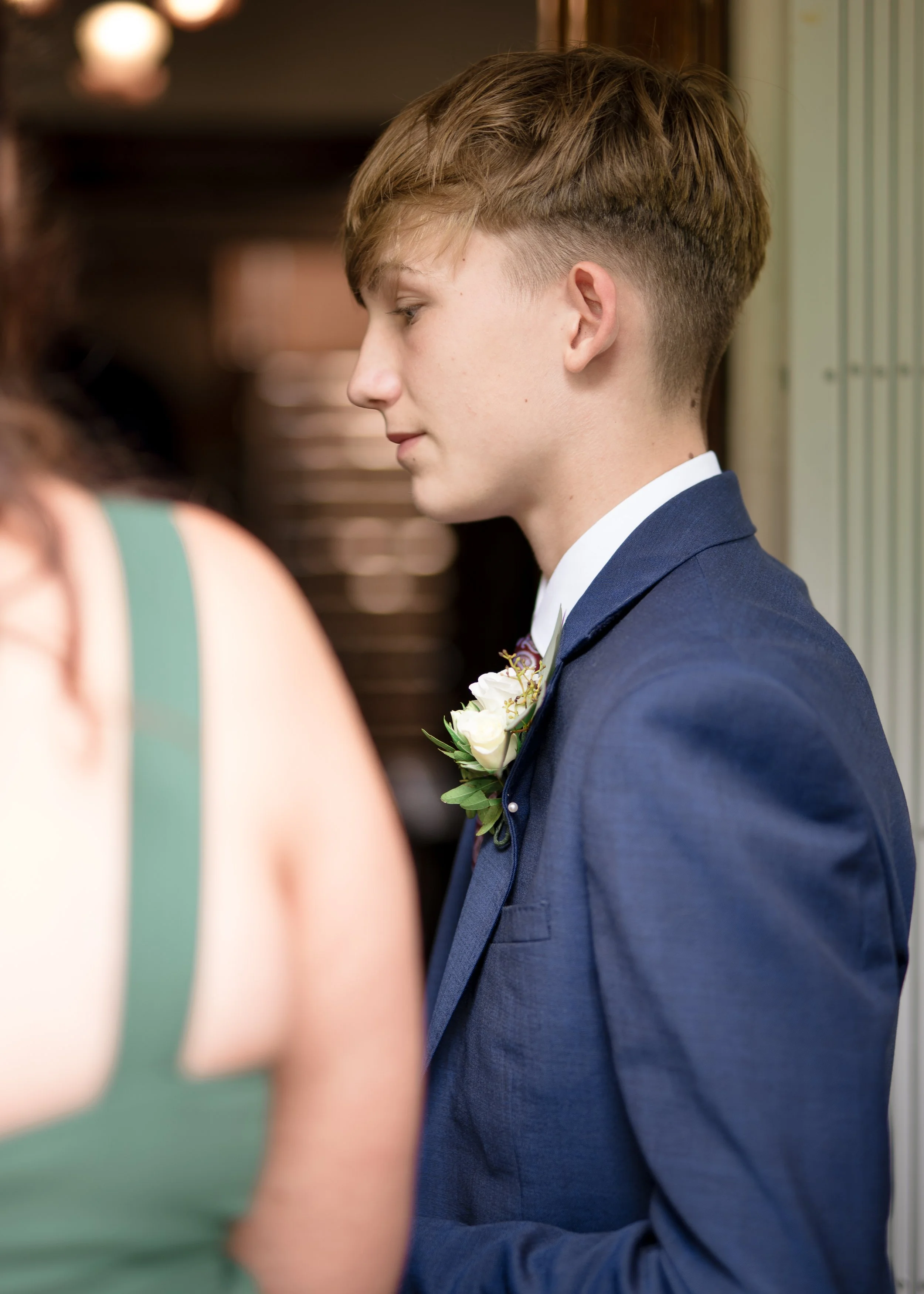 A young man in a blue suit with a white shirt and boutonniere stands with his eyes closed, appearing to be in a moment of reflection or prayer, near a woman whose shoulder is partially visible in the foreground.