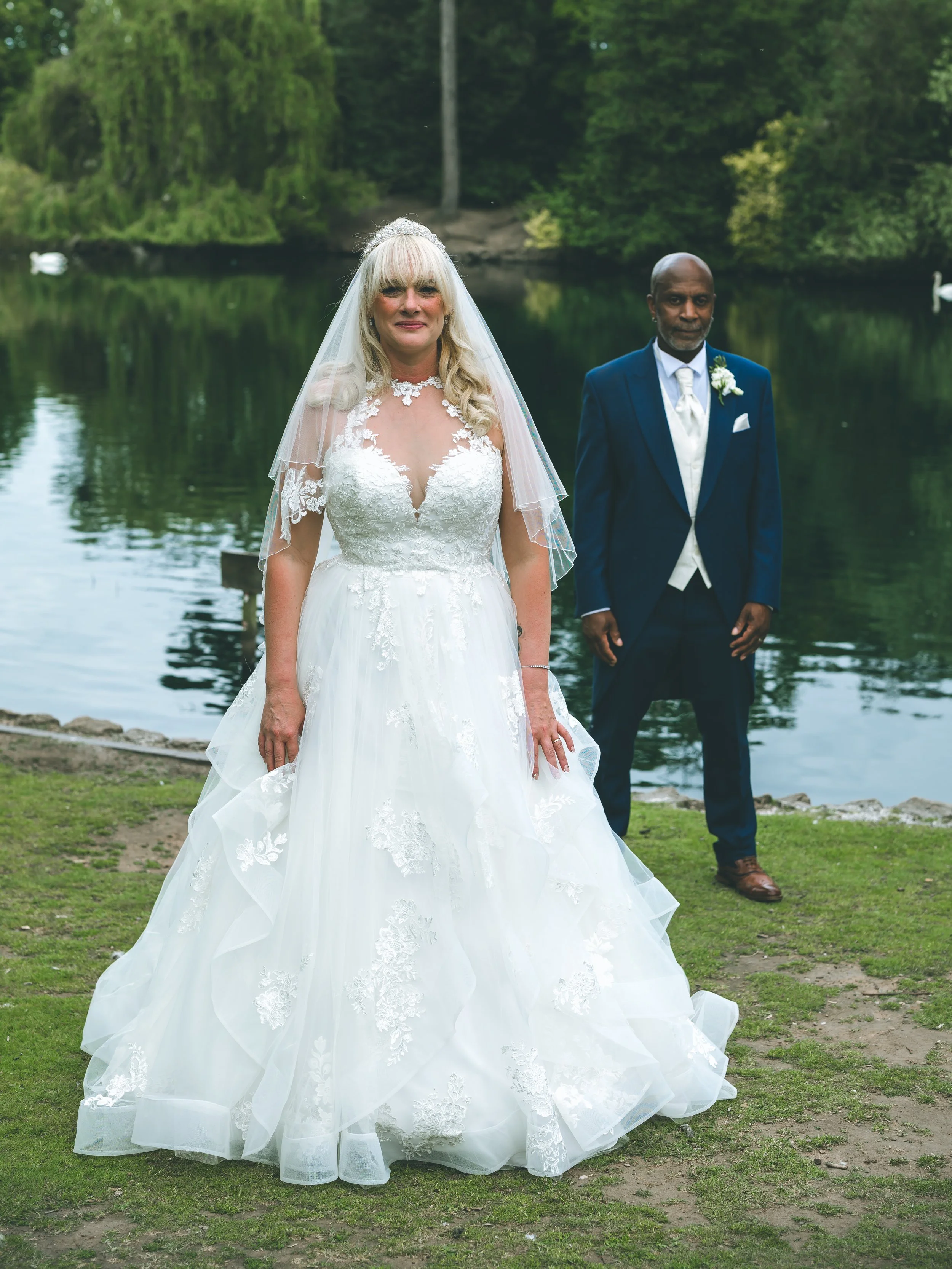 A bride with long blonde hair wearing a white wedding dress with lace details and a veil, standing outdoors by a lake, with a groom dressed in a navy blue suit and white shirt standing behind her.