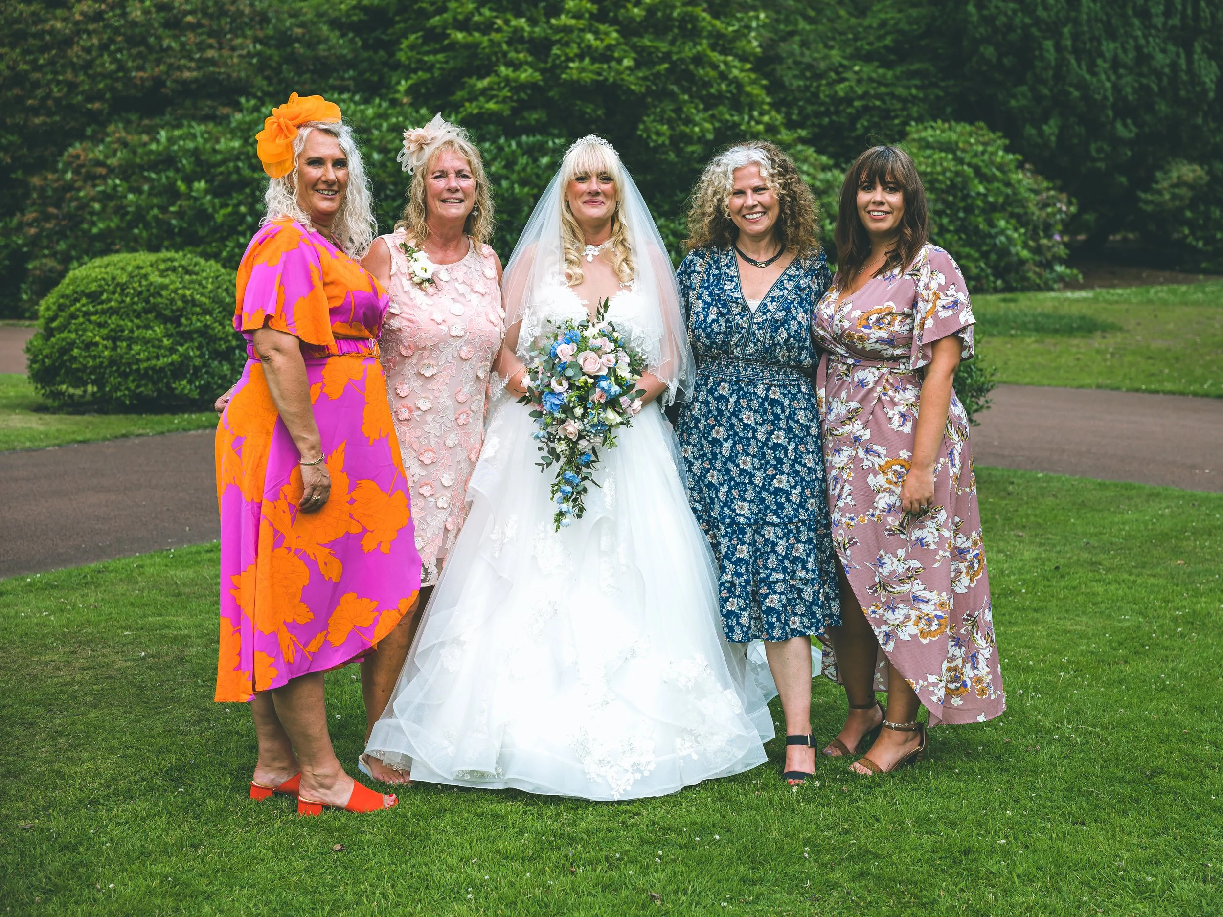A group of five women outdoors on a grassy lawn, with trees and bushes in the background. One woman is in a white wedding dress holding a bouquet, and the others are dressed in colorful dresses. They are all smiling and standing close together.