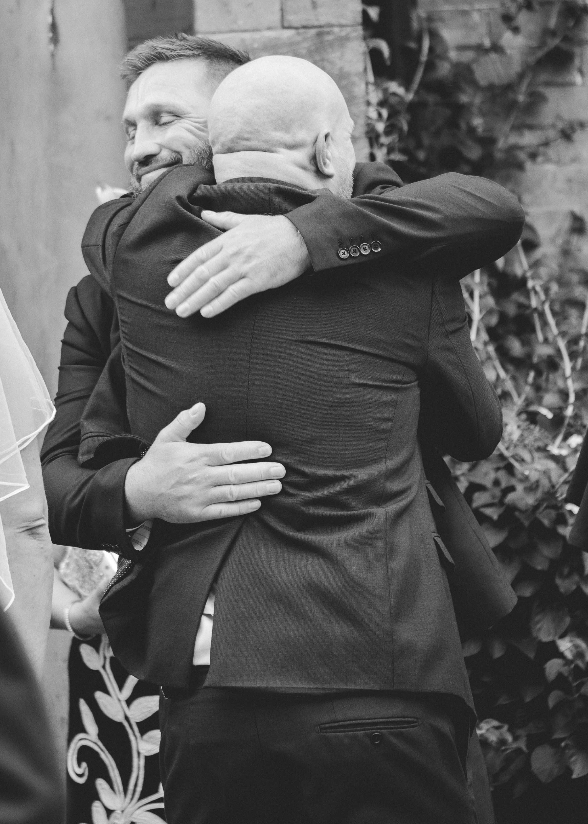 Two men hugging each other, one with a beard and the other bald, both dressed in suits. The man with a beard is smiling with eyes closed. The background includes a wall with plants.