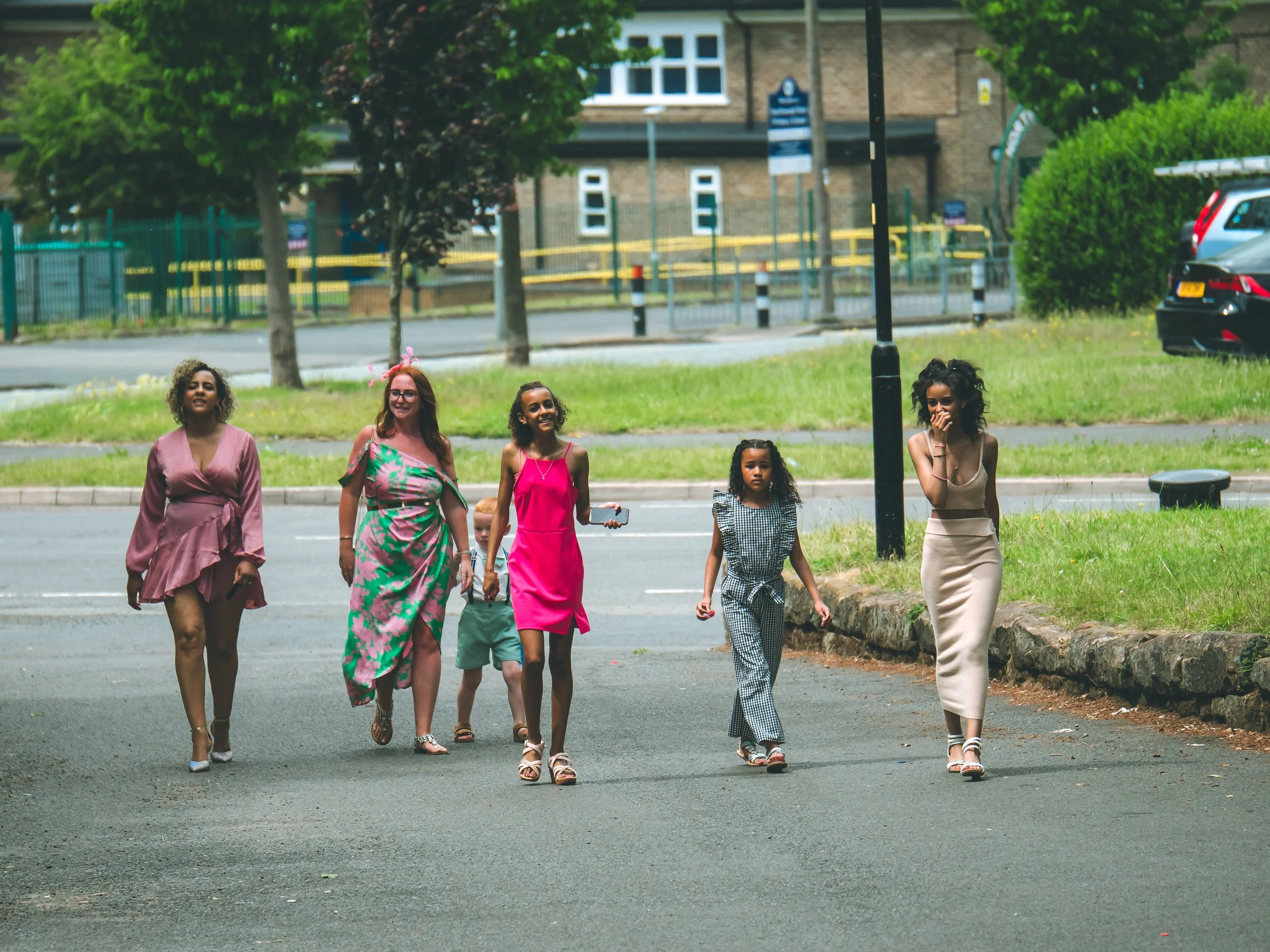 A diverse group of six women and children walking on a street sidewalk, smiling and enjoying a sunny day, with trees, parked cars, and a brick building in the background.