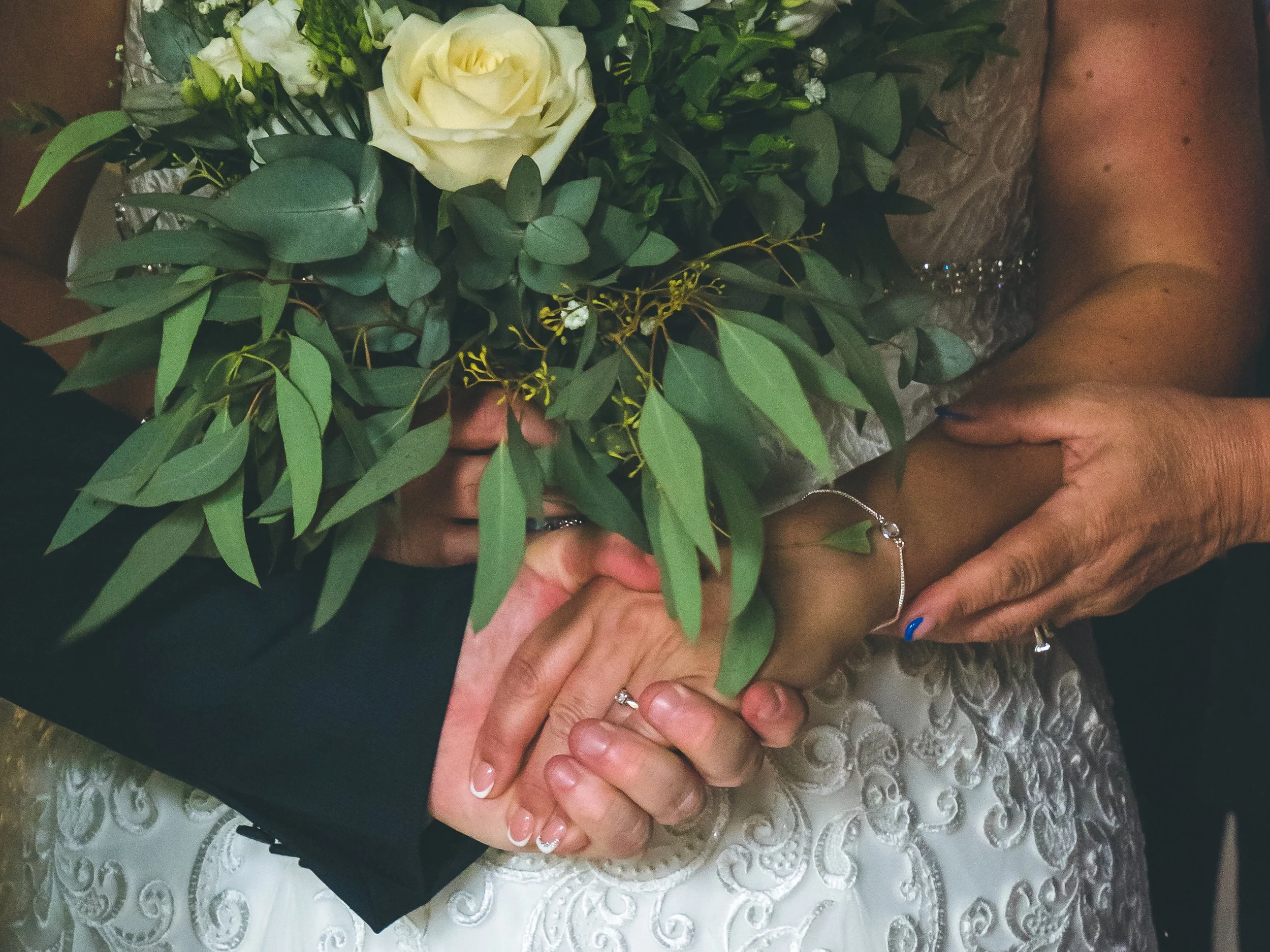 Close-up of a bouquet of white roses and green foliage being held by two people, one with painted nails and the other with a silver bracelet.