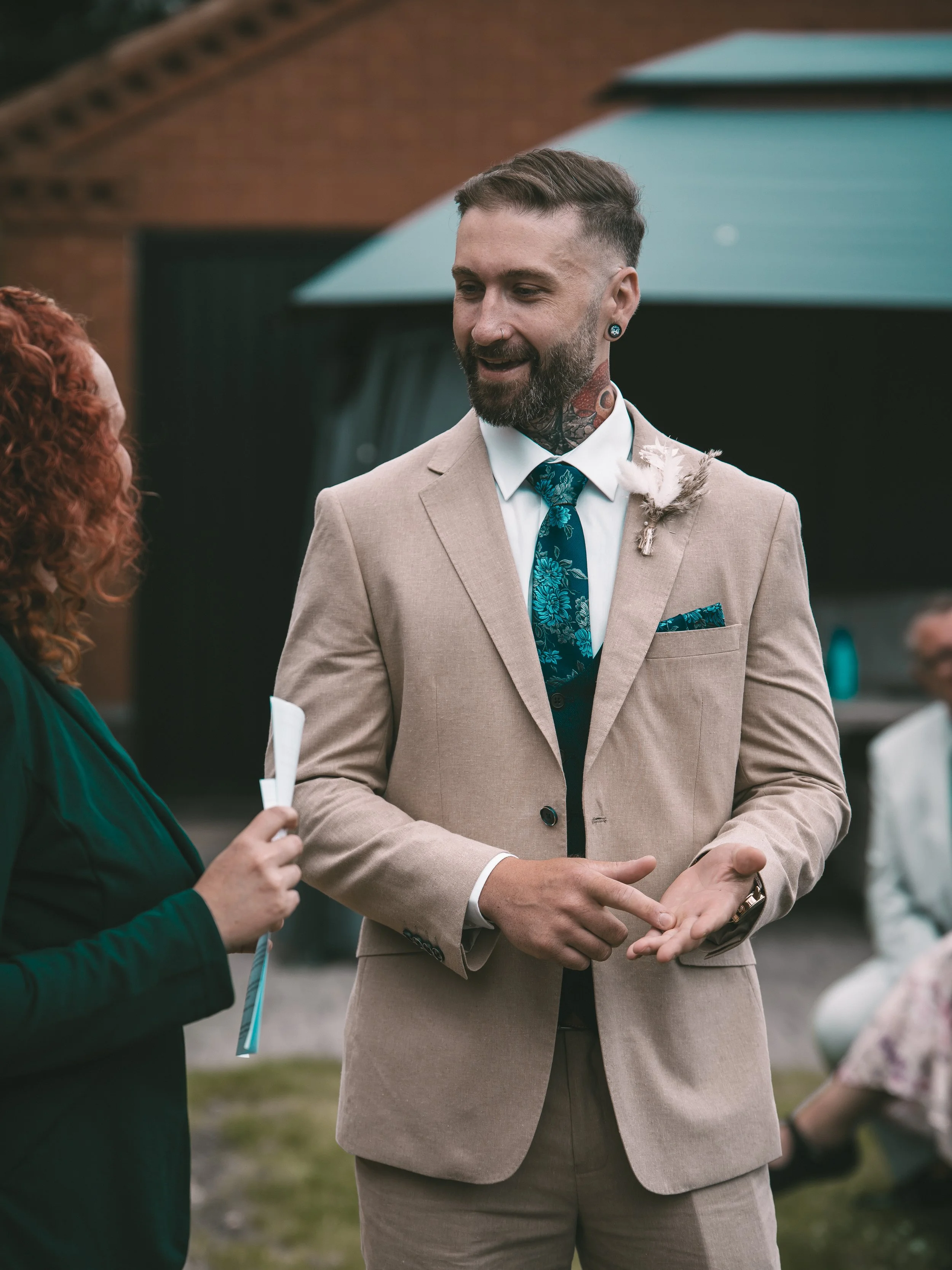 A man in a beige suit and tie with a floral pattern, with a white boutonniere, is smiling and talking to a woman with curly red hair, who is holding a paper or program. The outdoor setting appears to be a social event or wedding.