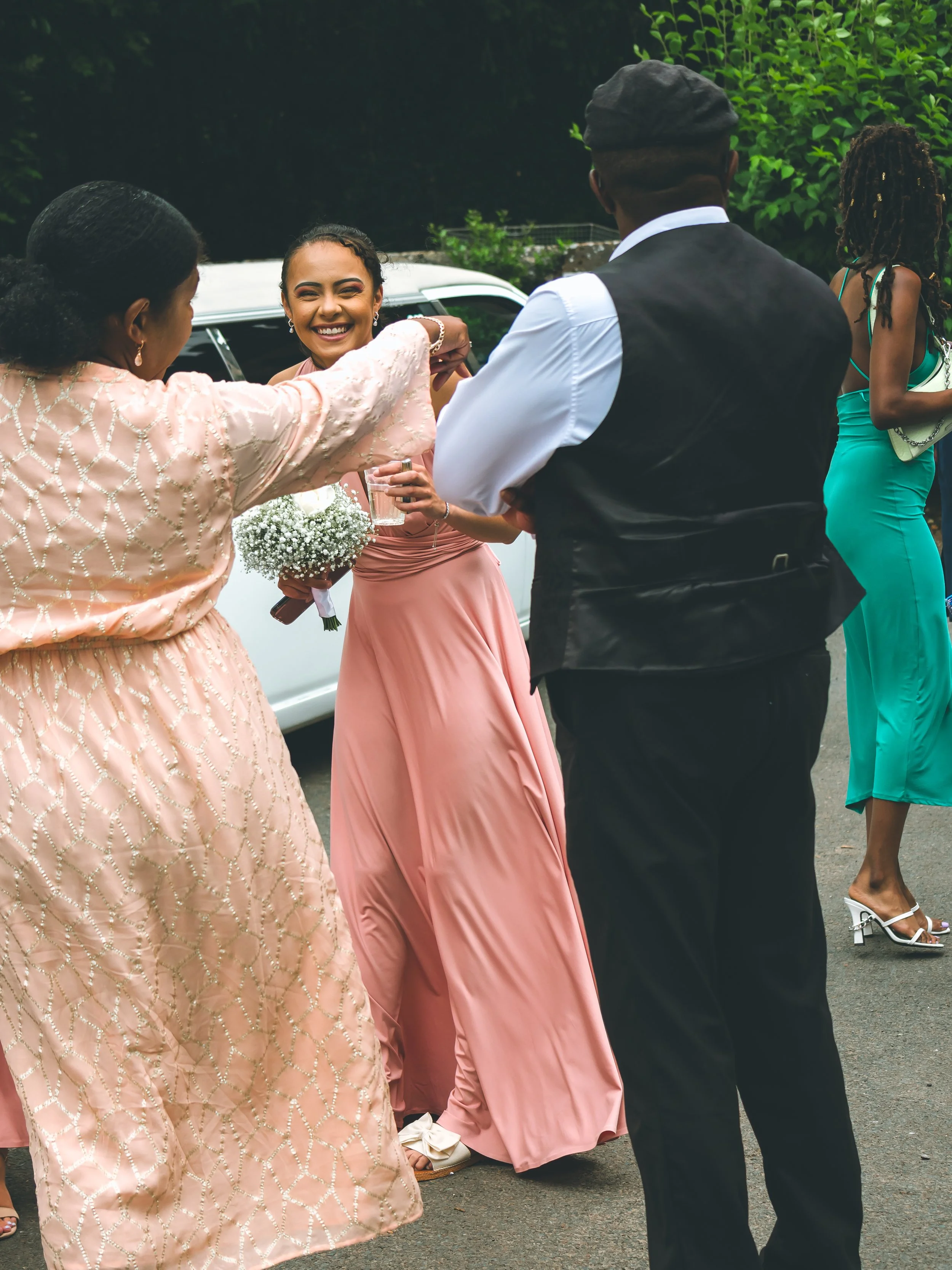 A group of people dressed in formal attire, standing outdoors near a white vintage car, with green trees in the background. One woman in a pink dress is smiling, holding a flower bouquet, and participating in a social gathering or celebration.