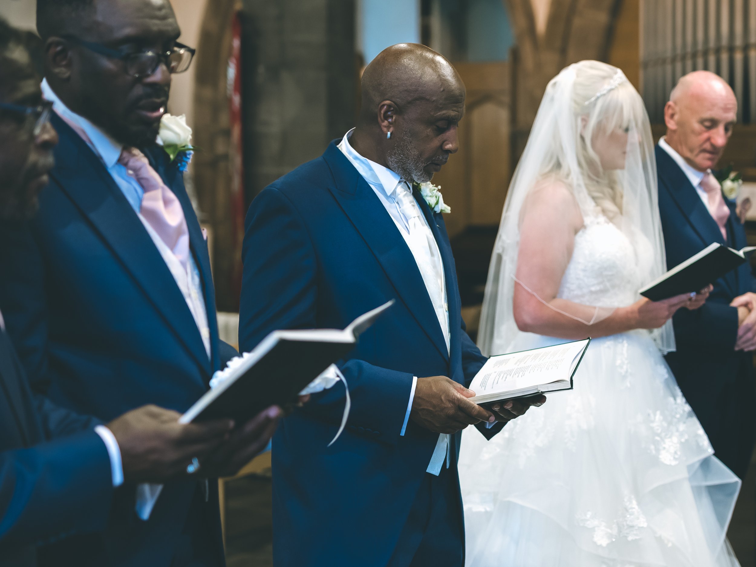 A group of five people dressed formally, standing in a row inside a church, reading from booklets during a wedding ceremony. The bride wears a white dress and veil, and all are holding open books, likely hymnals or prayer books.