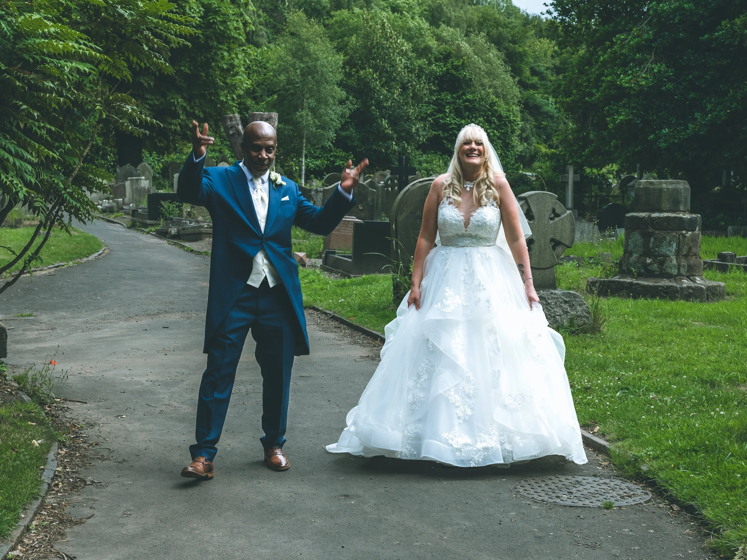 A wedding couple walking through a cemetery surrounded by lush green trees and grave markers. The bride is wearing a white wedding dress and the groom is wearing a blue suit.