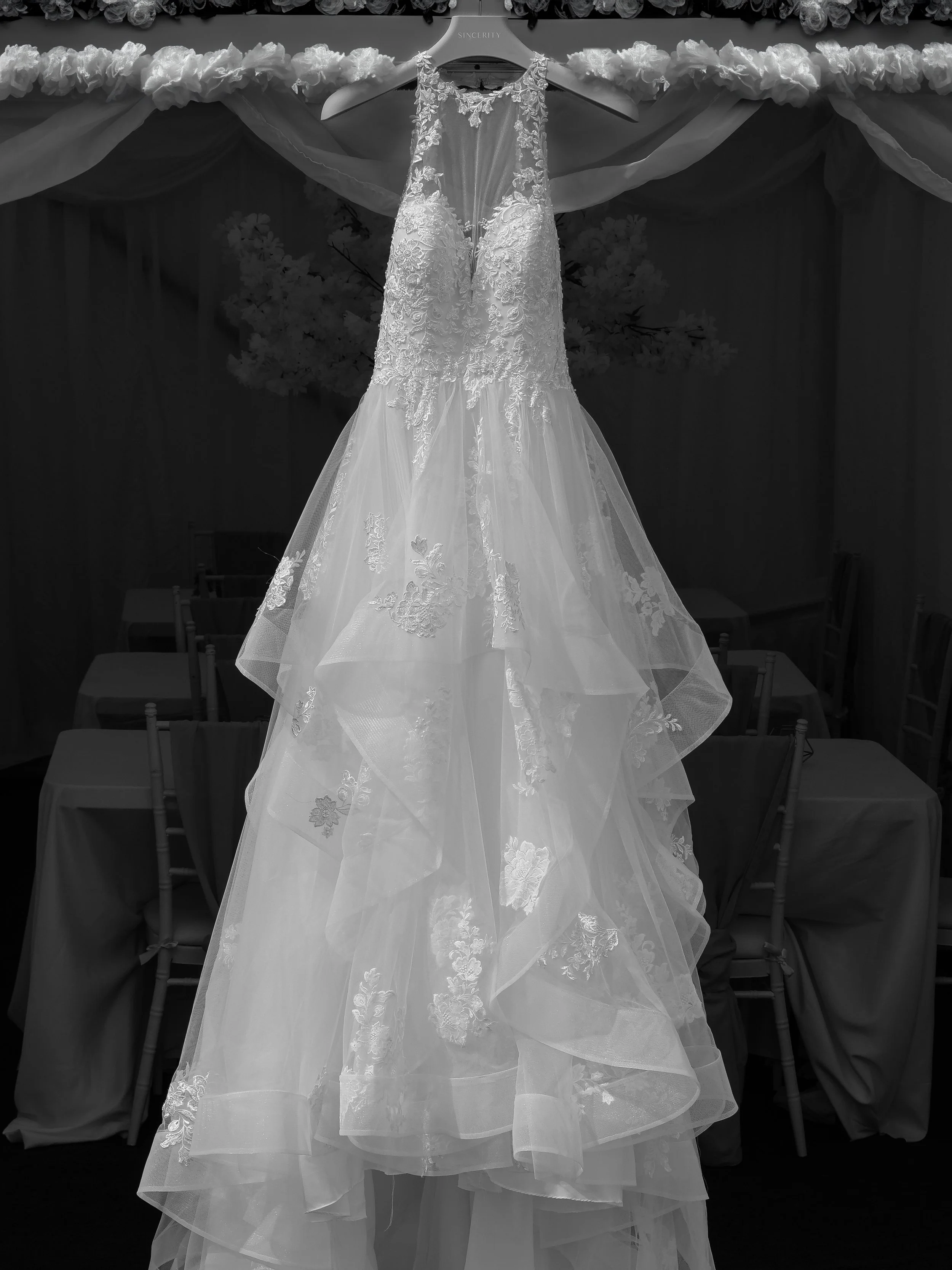 A wedding dress with lace detailing hanging on a hanger, decorated with flowers, against a dark background.