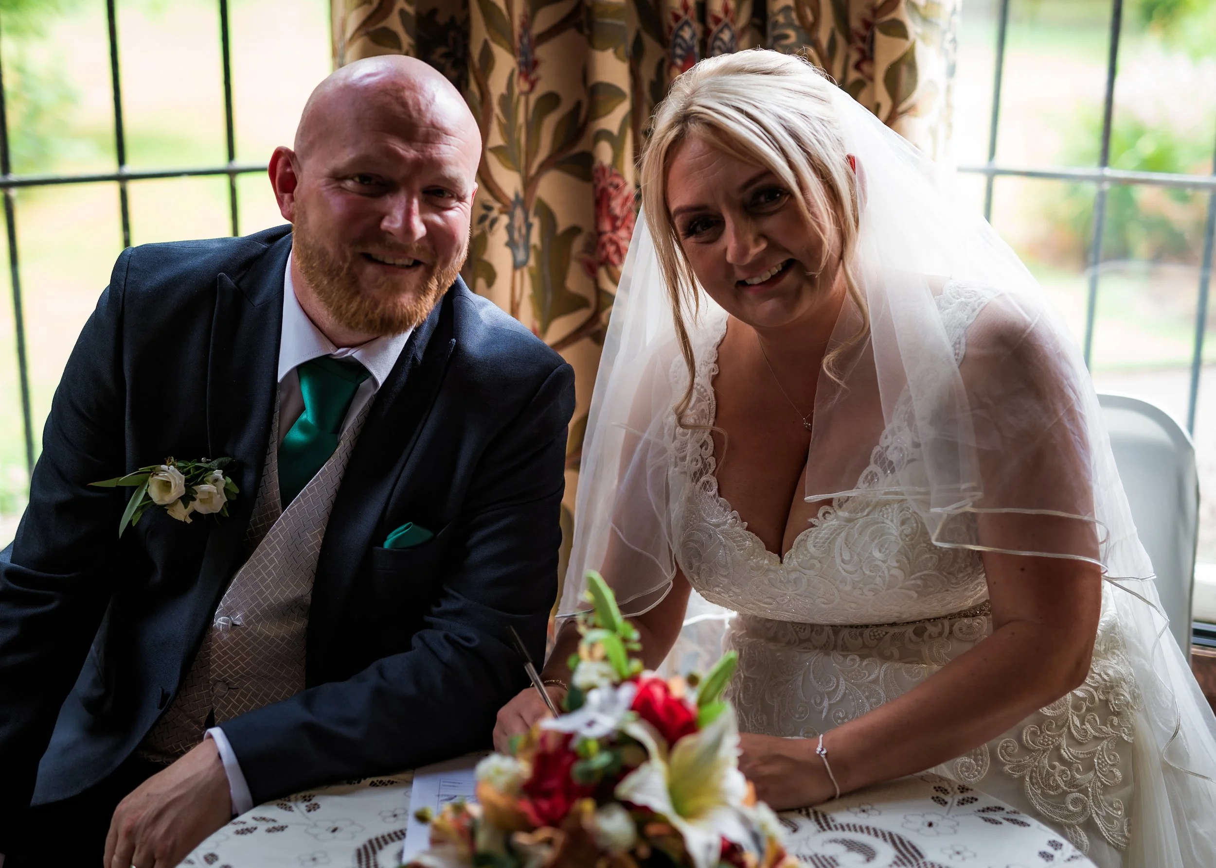 A bride and groom are signing documents at their wedding ceremony, with a bouquet of flowers on the table in front of them.