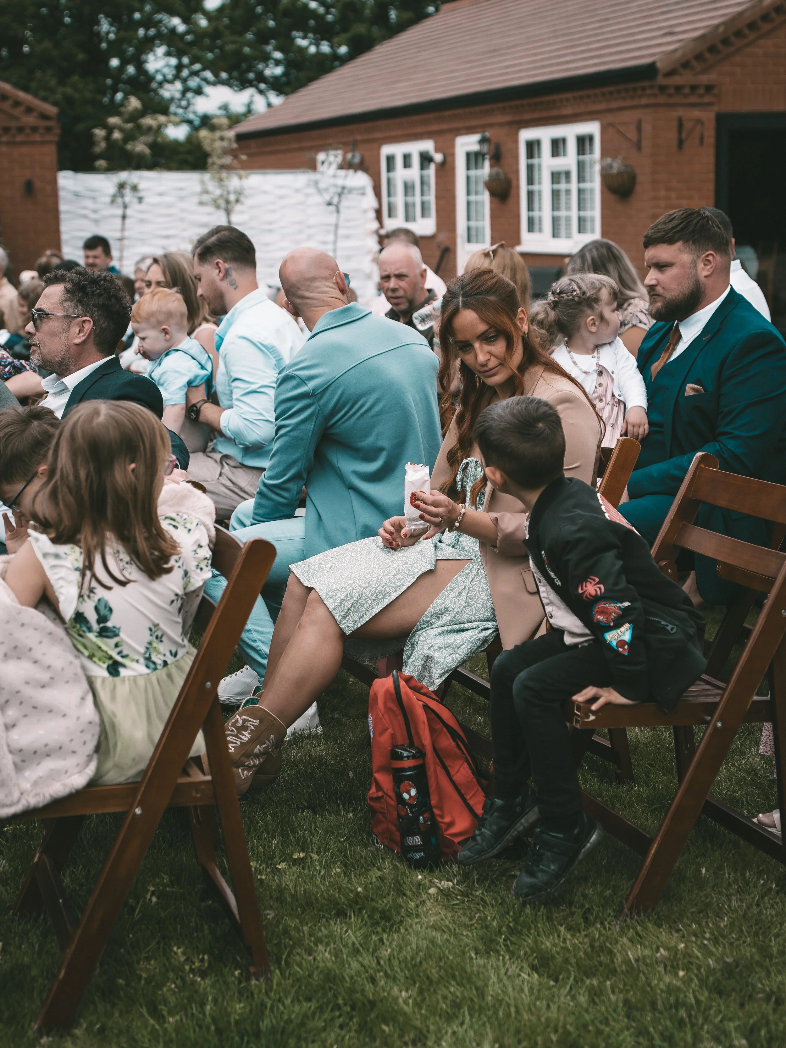 A group of people, including children and adults, seated outdoors on wooden chairs during daytime. They are in front of brick houses with white-framed windows. The scene suggests a social gathering or event.
