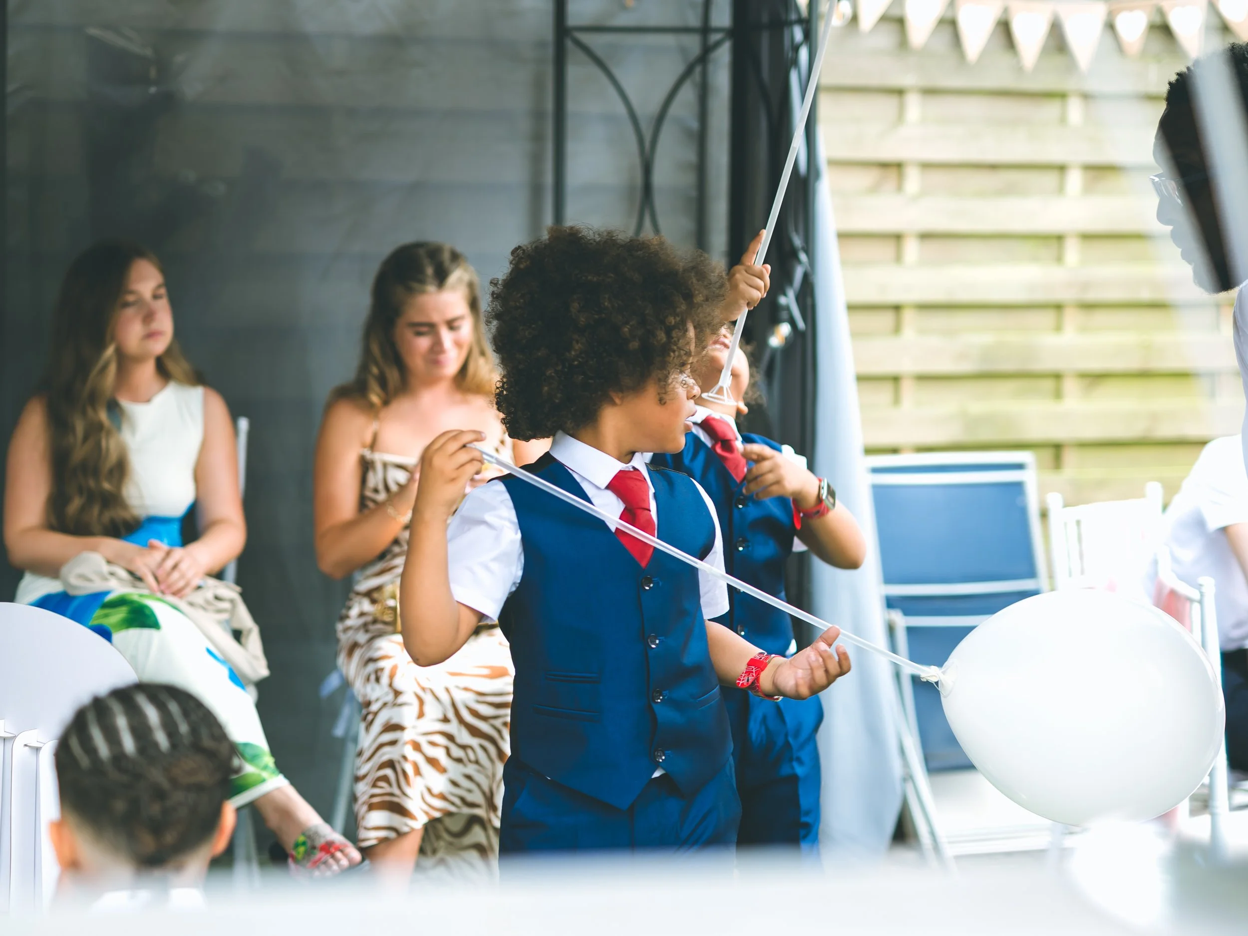 Group of children at a celebration, some holding white balloons and one holding a balloon and a string, with one child in a school uniform and others in casual clothes, outdoors near a black tent and a wooden fence.