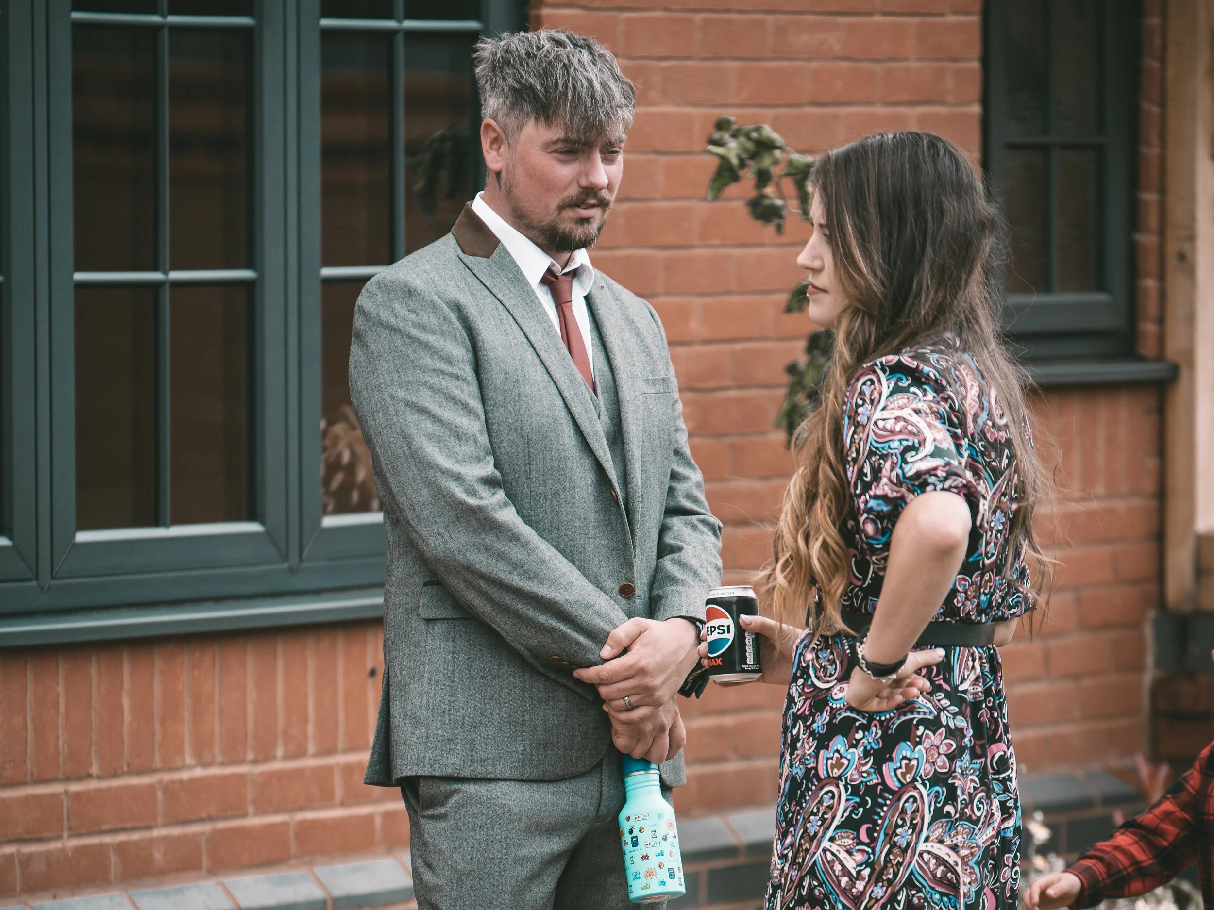 A man and a woman stand close to each other outside, facing each other with their hands together, with a brick wall and window behind them. The man is dressed in a gray suit with a red tie and the woman is wearing a colorful, patterned dress. The wom