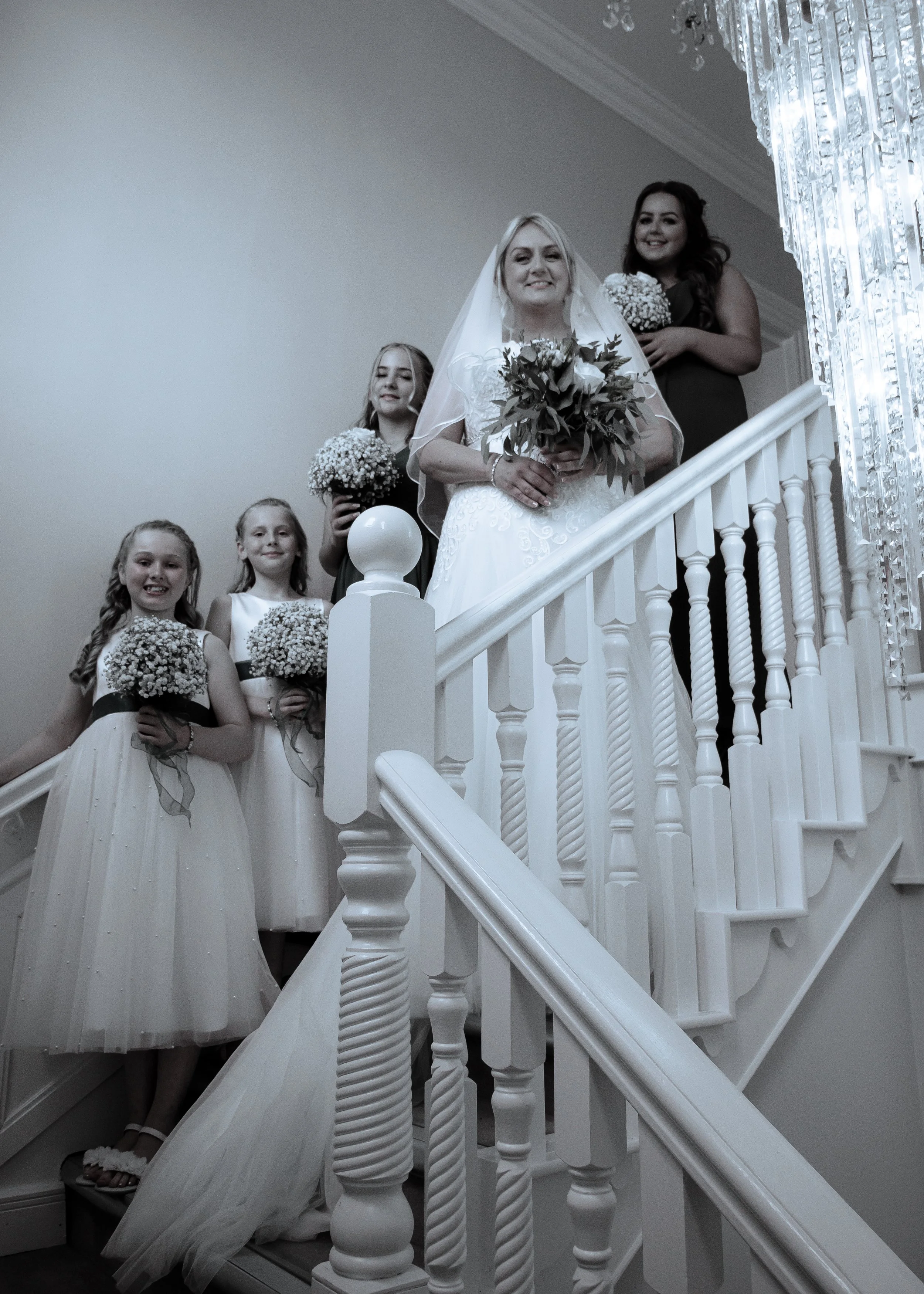 A bride and two bridesmaids on a staircase, with three young flower girls standing below, all holding bouquets. The bride is in a white wedding dress and lace veil, smiling at the camera, while the bridesmaids and flower girls are smiling and looking