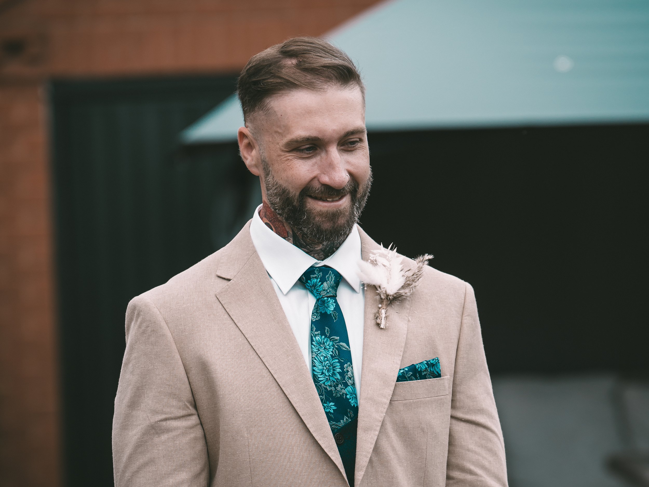 A man with a beard and short hair, dressed in a beige suit with a blue floral tie and pocket square, smiling outdoors.
