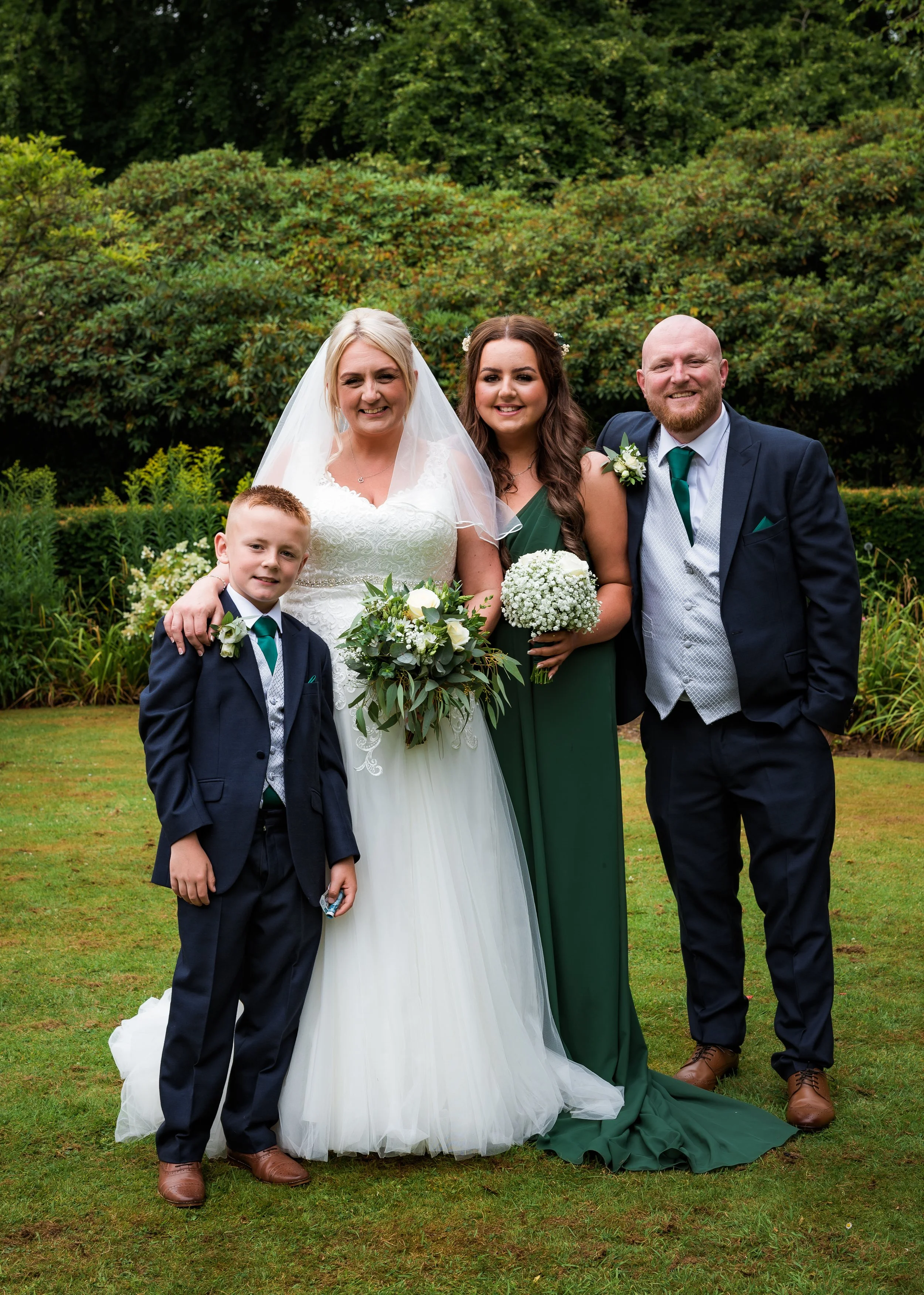 A wedding party group photo features a bride in a white gown and veil, holding a bouquet, standing next to a woman in a green dress, and a man in a dark suit with a green tie, all smiling outdoors with greenery in the background. A young boy in a sui