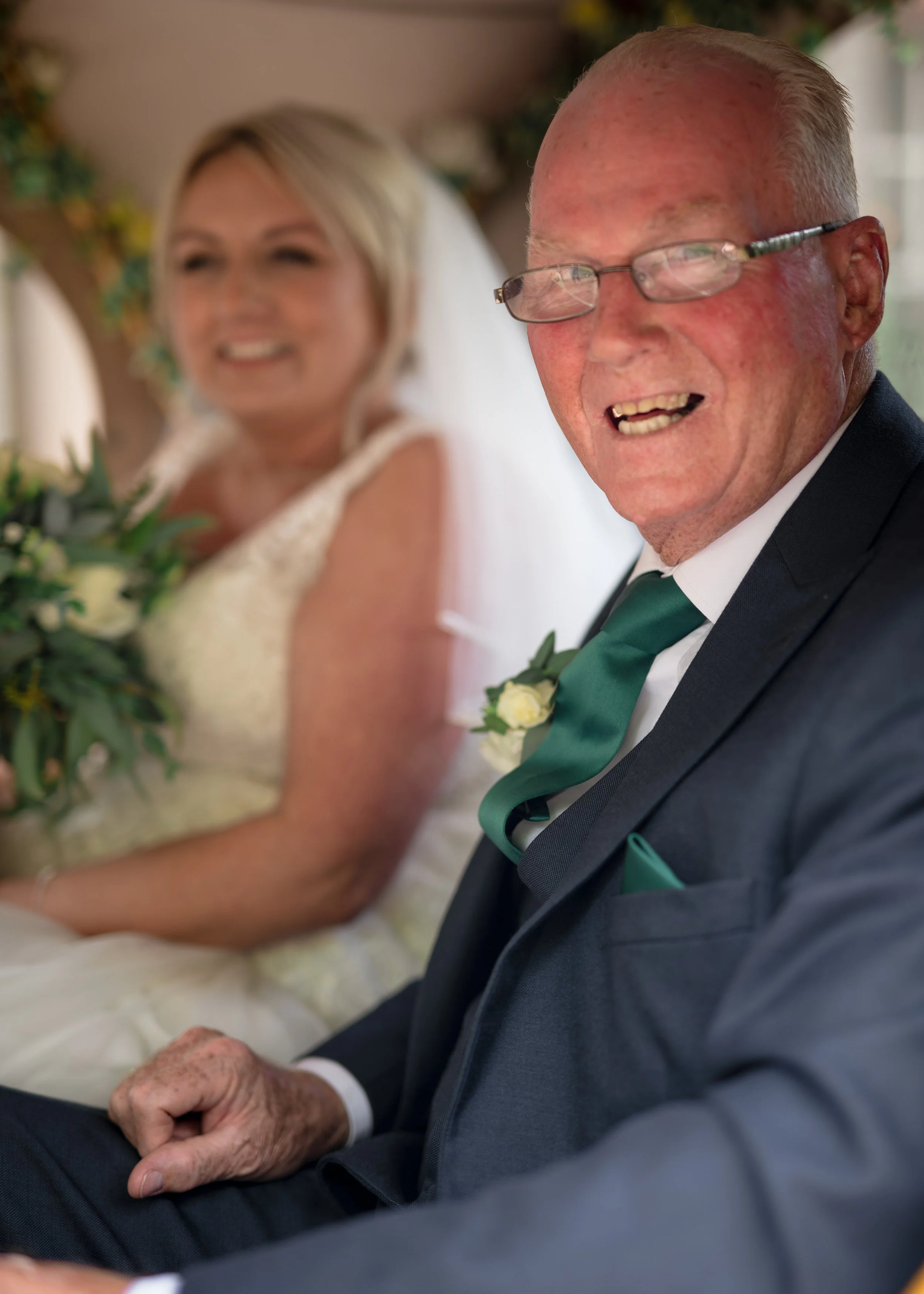 An elderly man dressed in a dark suit, with glasses and a green tie, sitting in a formal event. Behind him, a woman in a white dress with short blonde hair is smiling, holding a bouquet of flowers. Both are smiling and appear to be at a wedding or ce