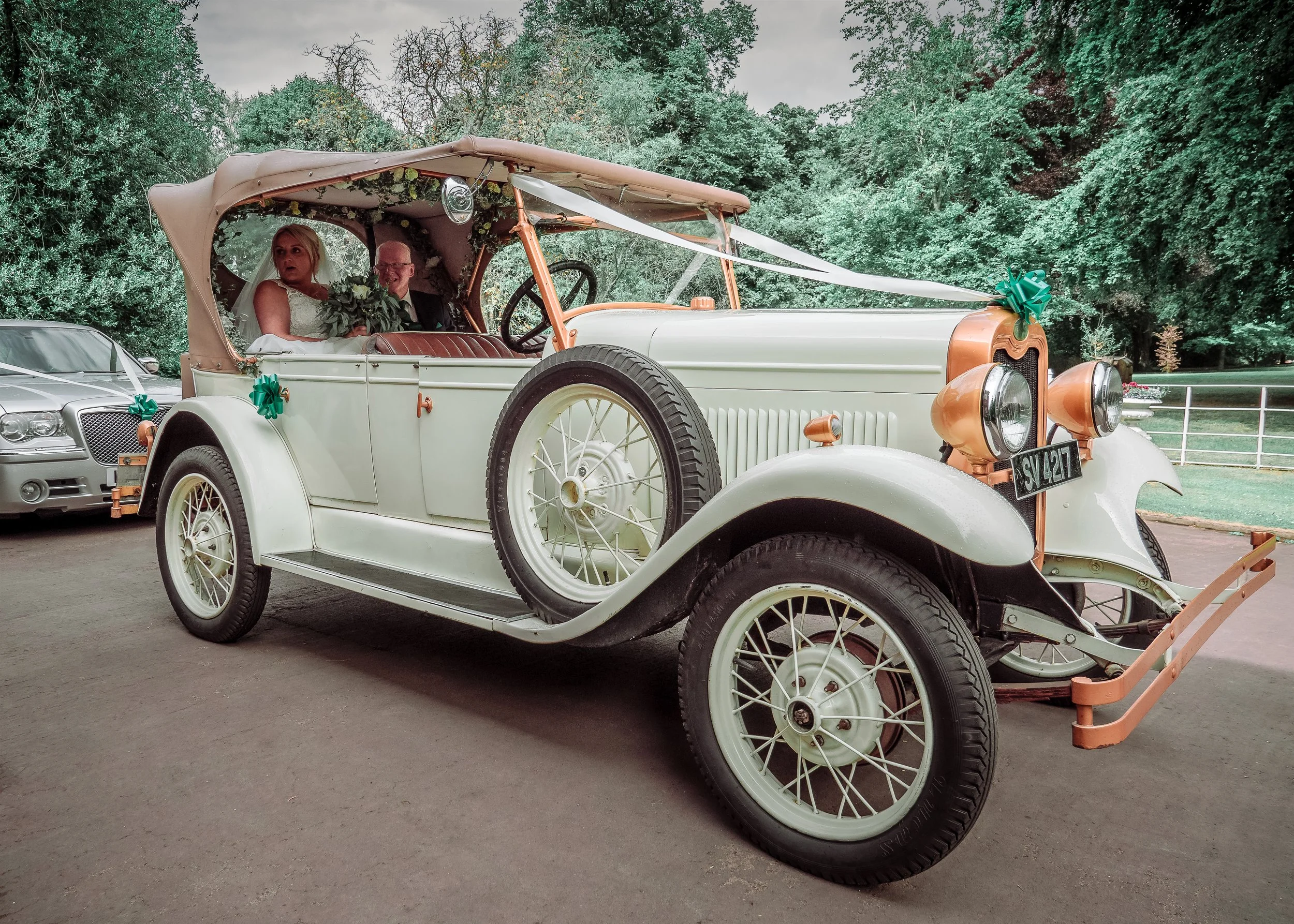 A vintage white wedding car decorated with green ribbons parked outdoors, with a bride and groom sitting inside.
