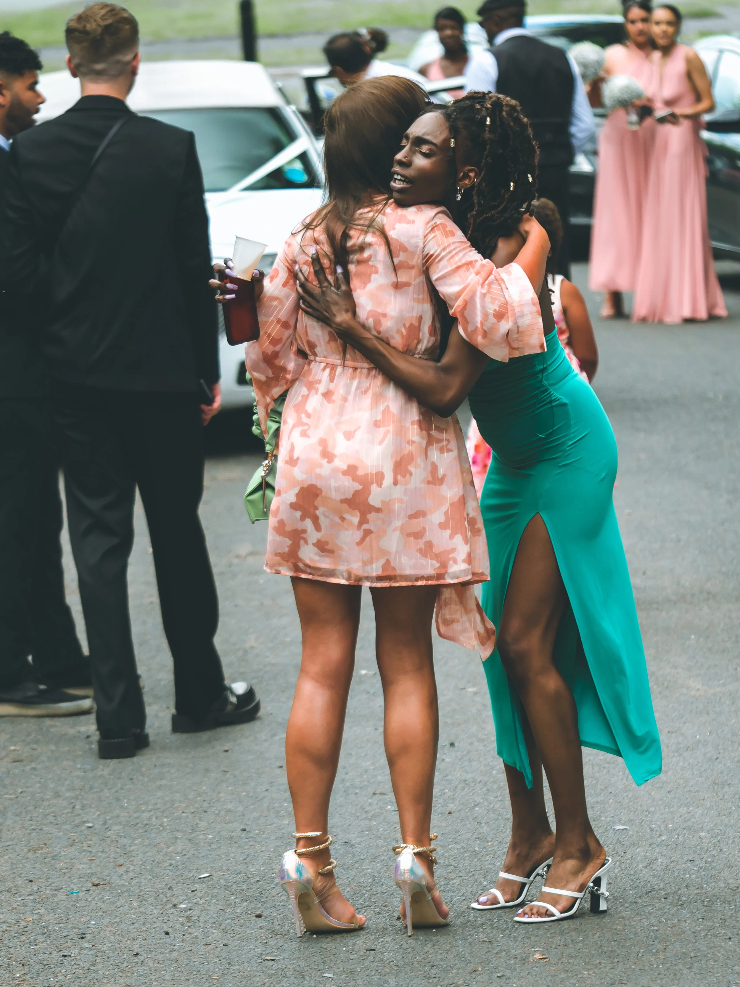 Two women hugging at an outdoor event, one wearing a peach camouflage dress and high heels, the other in a teal dress with high-heeled sandals. Several people in formal attire are in the background near cars.