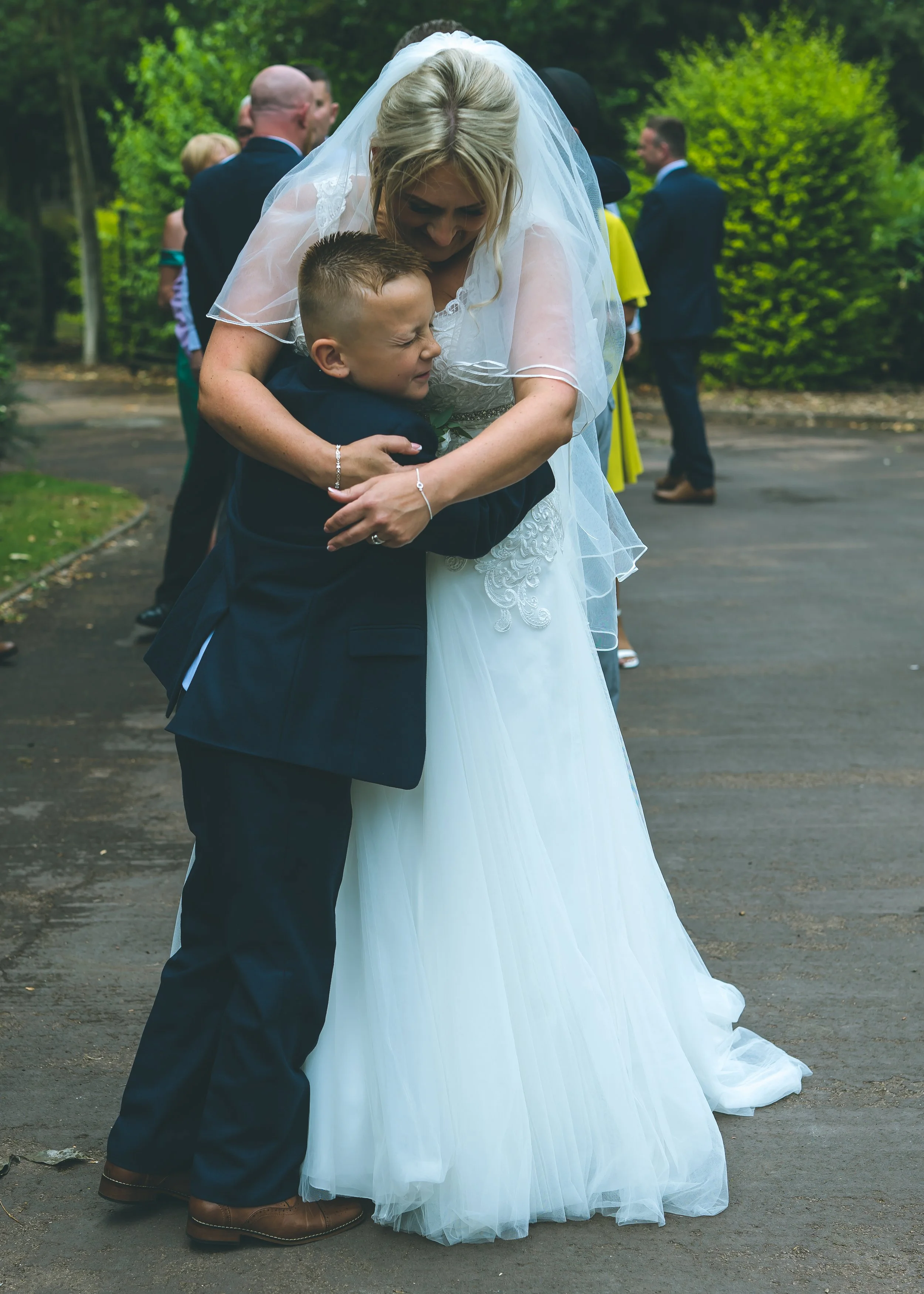 A woman dressed in a white wedding gown with a veil gently embracing a young boy in a dark suit, outside among trees and other well-dressed people, at a wedding ceremony.