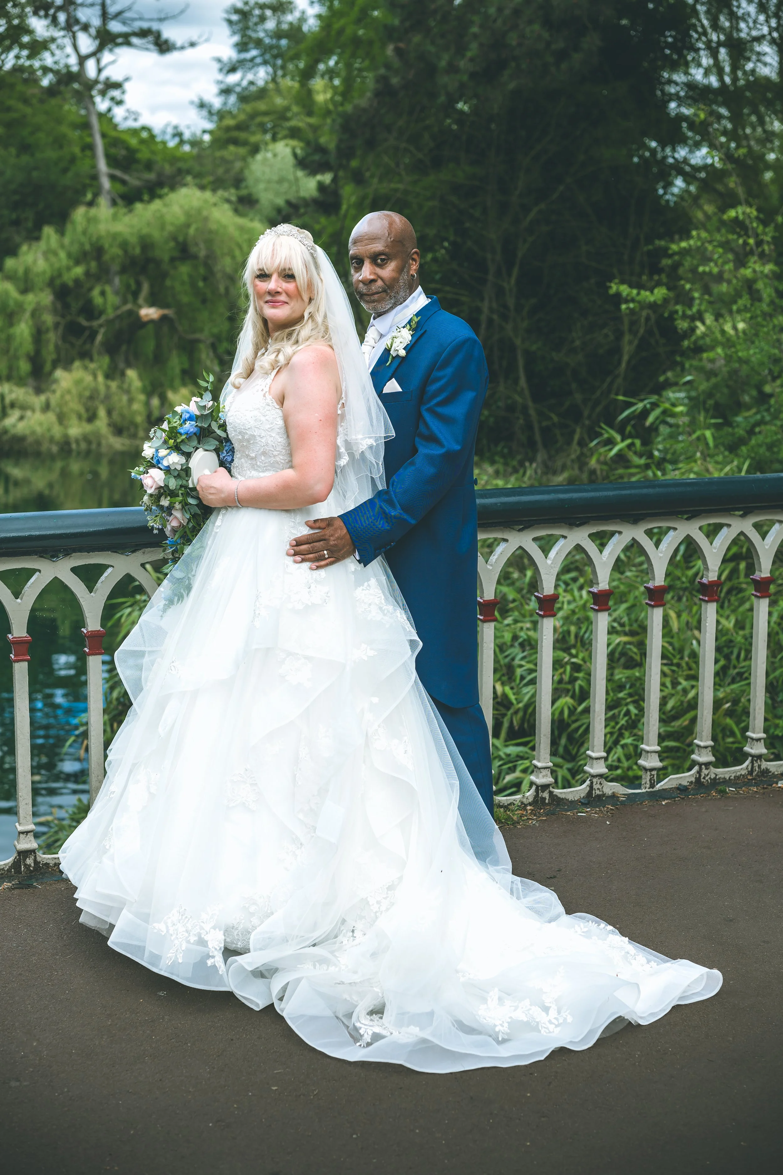 A bride in a white wedding dress with a long train and lace details, holding a bouquet, and a groom in a blue suit with a white shirt, standing outdoors on a bridge near a body of water and surrounded by green trees.