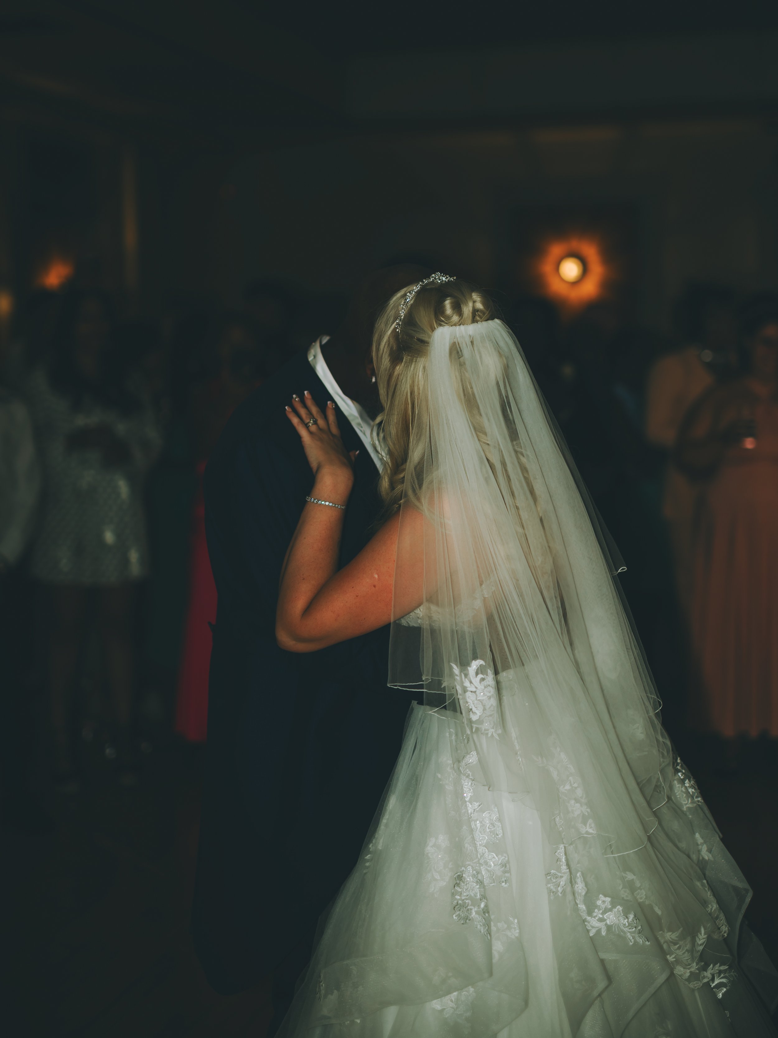 A bride and groom sharing a kiss at their wedding reception, with the bride wearing a white wedding gown and veil, and the groom dressed in a black tuxedo. The background is dimly lit with soft lighting and guests are visible at the tables.