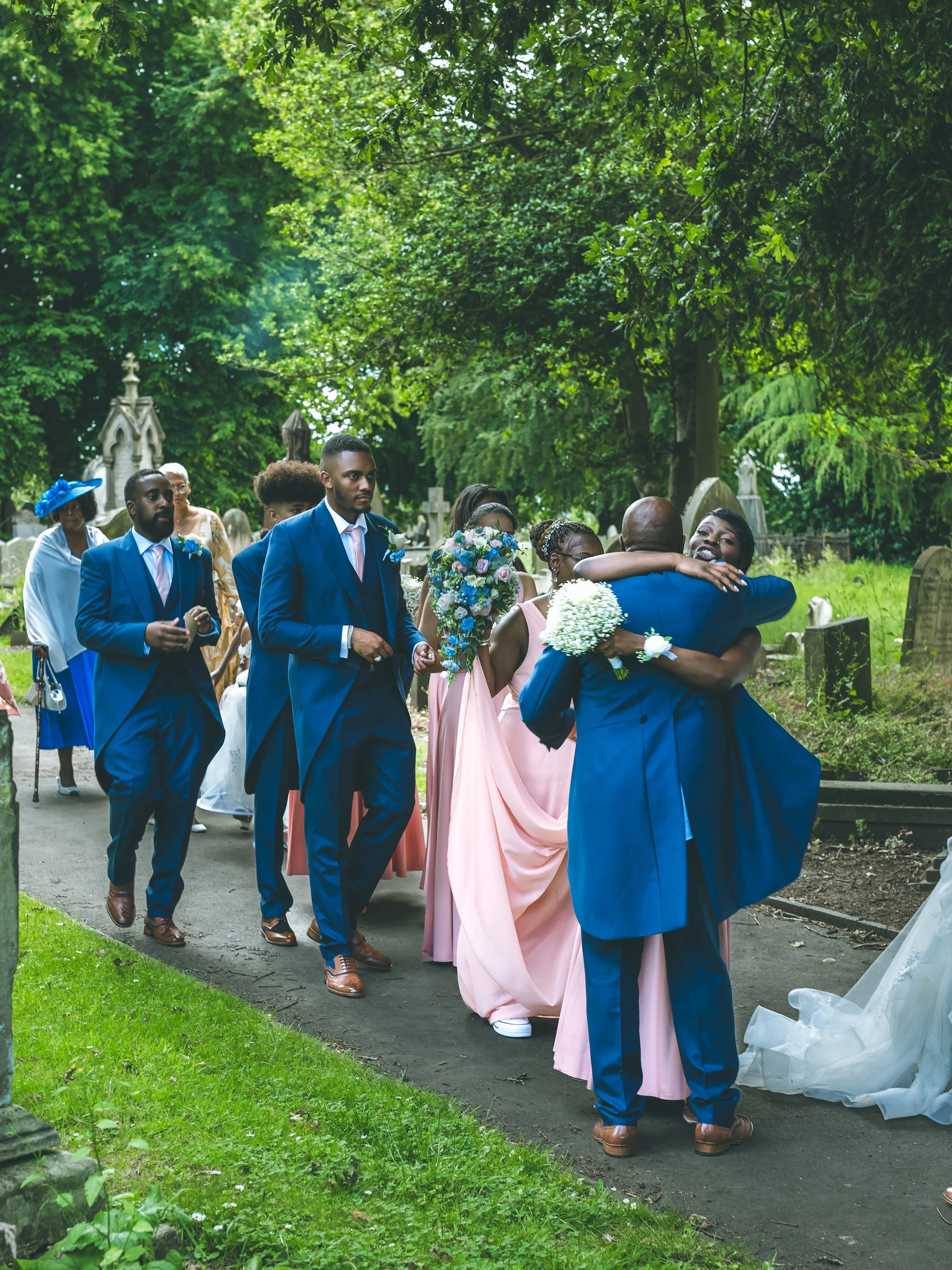 A group of people at a funeral or memorial service in a cemetery, with some hugging and others holding flowers, surrounded by greenery and tombstones.