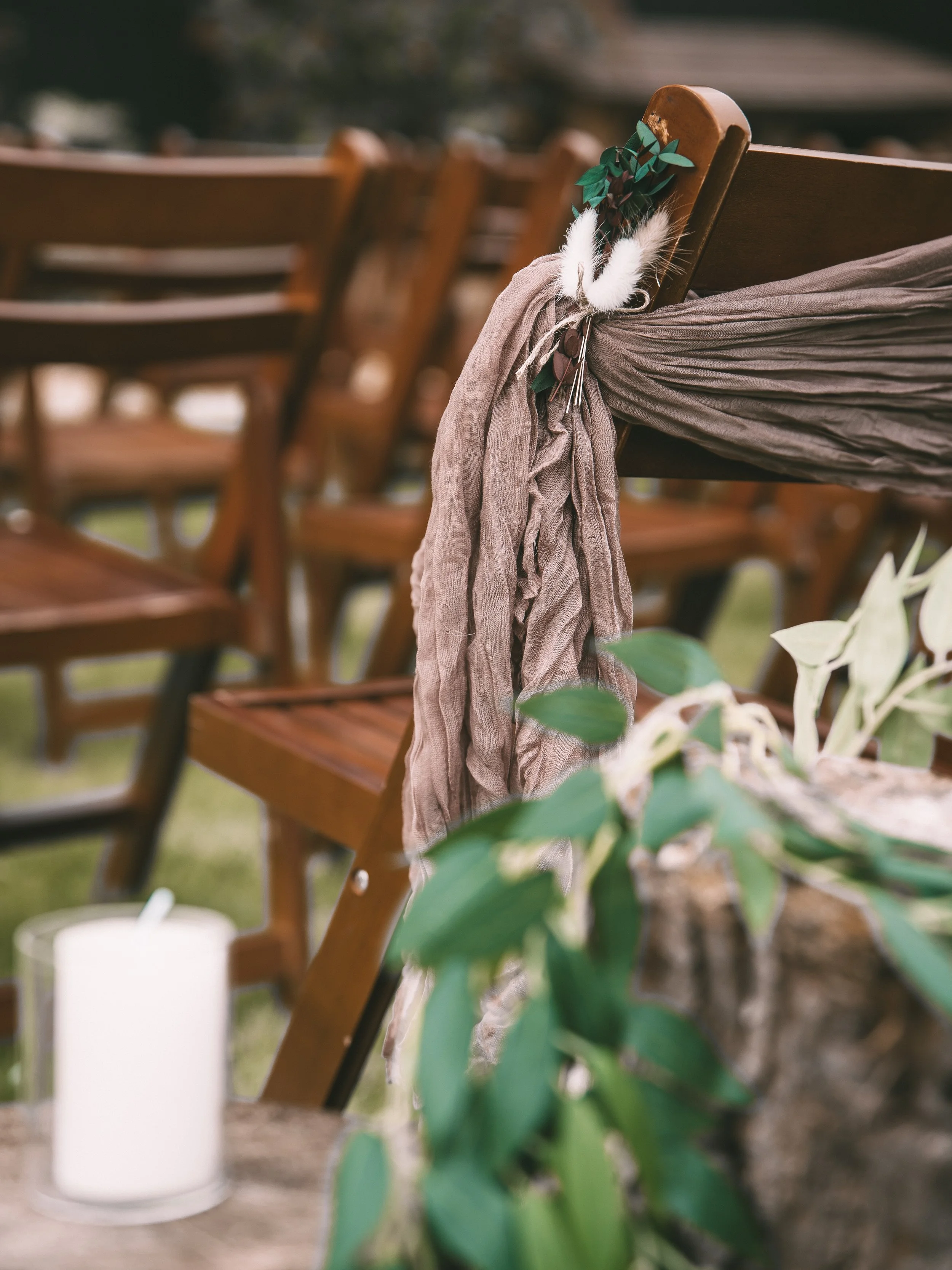 Wooden chairs decorated with floral and ribbon arrangements at an outdoor event, with a white candle in the foreground.