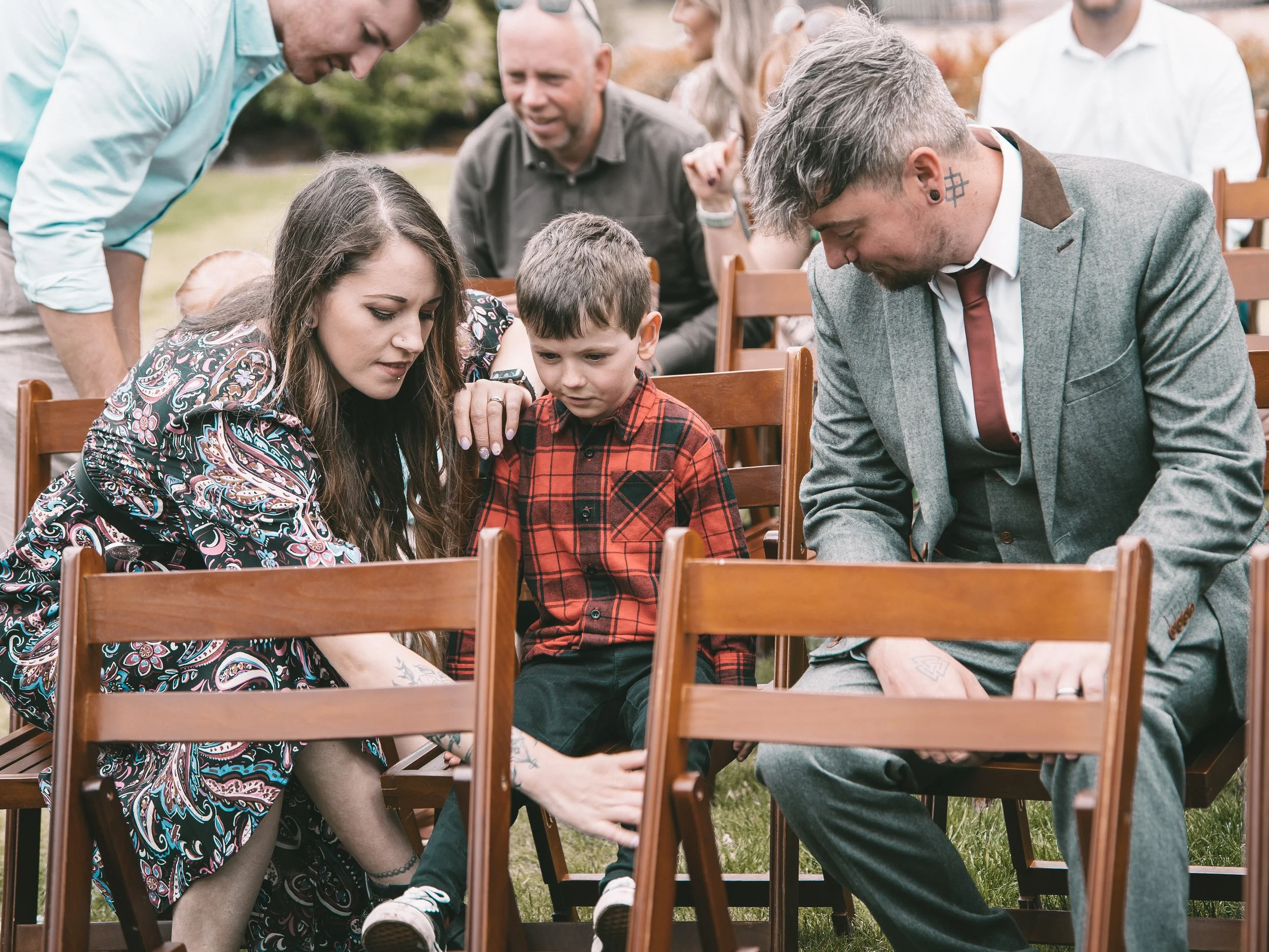 A group of people, including a woman, a young boy, and a man with tattoos, are gathered outdoors around a table, engaging in a shared activity.