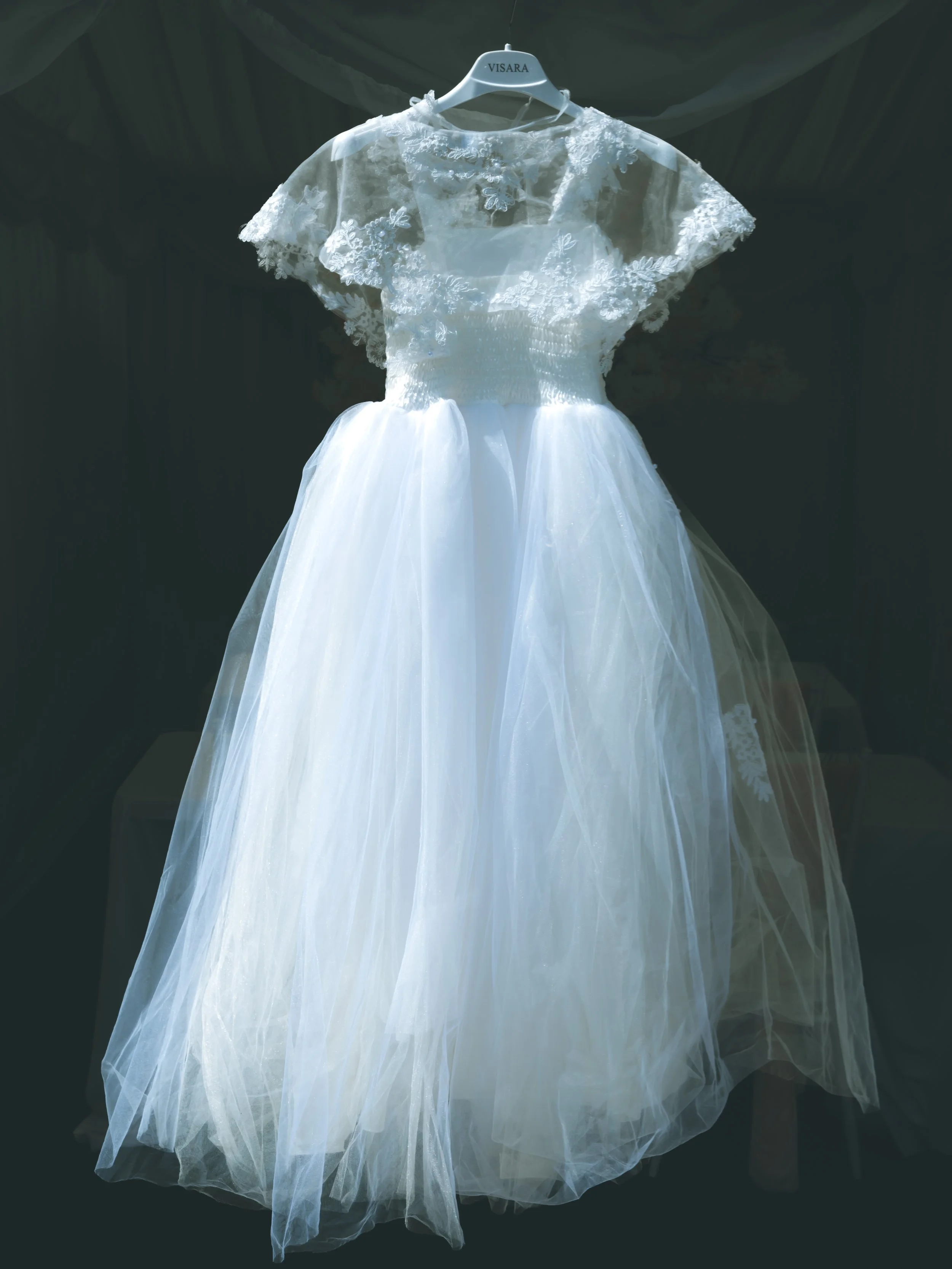 A white lace wedding dress with floral embroidery hanging on a hanger labeled 'VISARA' against a dark background.