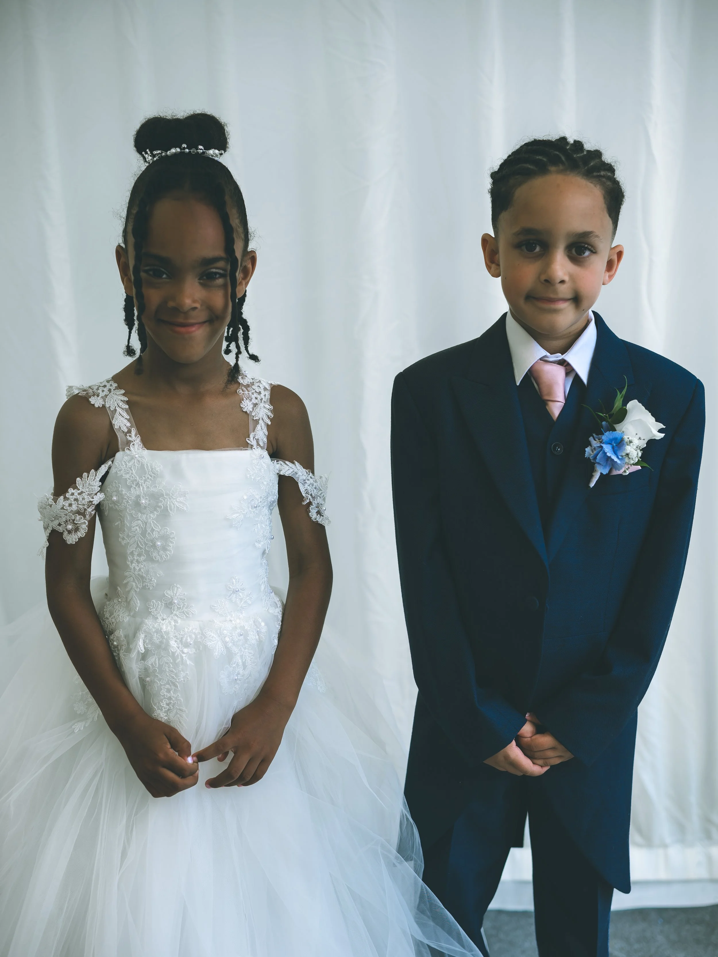 A young girl in a white lace dress and a boy in a navy blue suit with a pink tie and white boutonniere, standing side by side against a plain white backdrop.