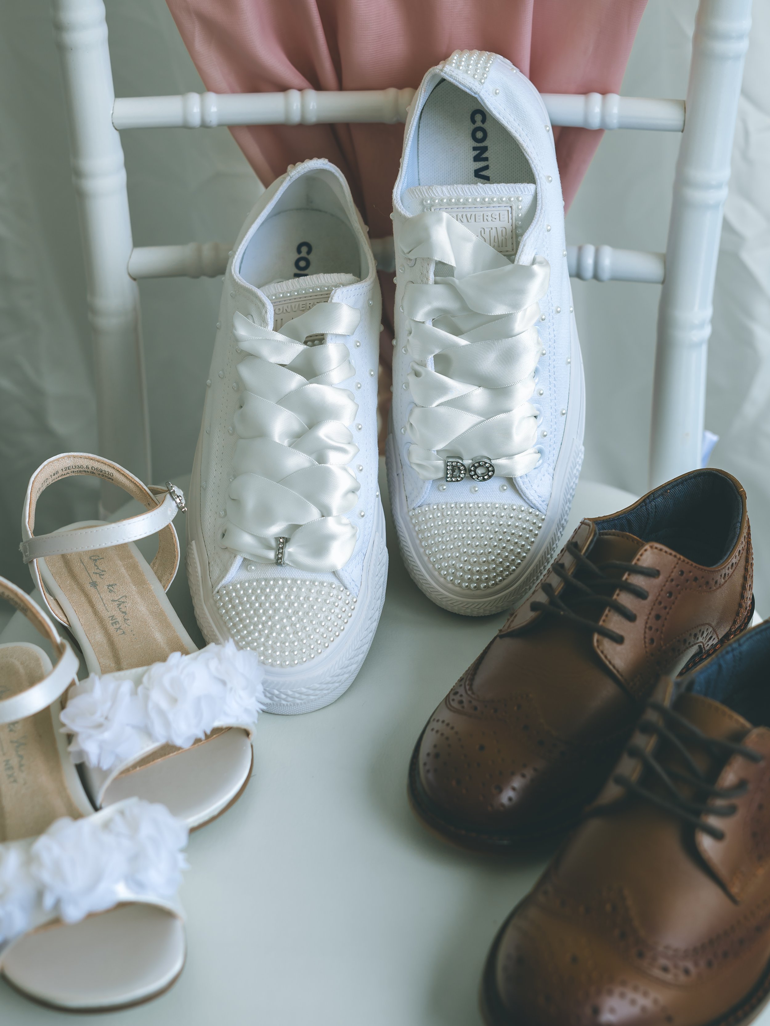Pair of white Converse sneakers with pearl embellishments and satin ribbons, placed on a surface with other shoes and a fabric background.