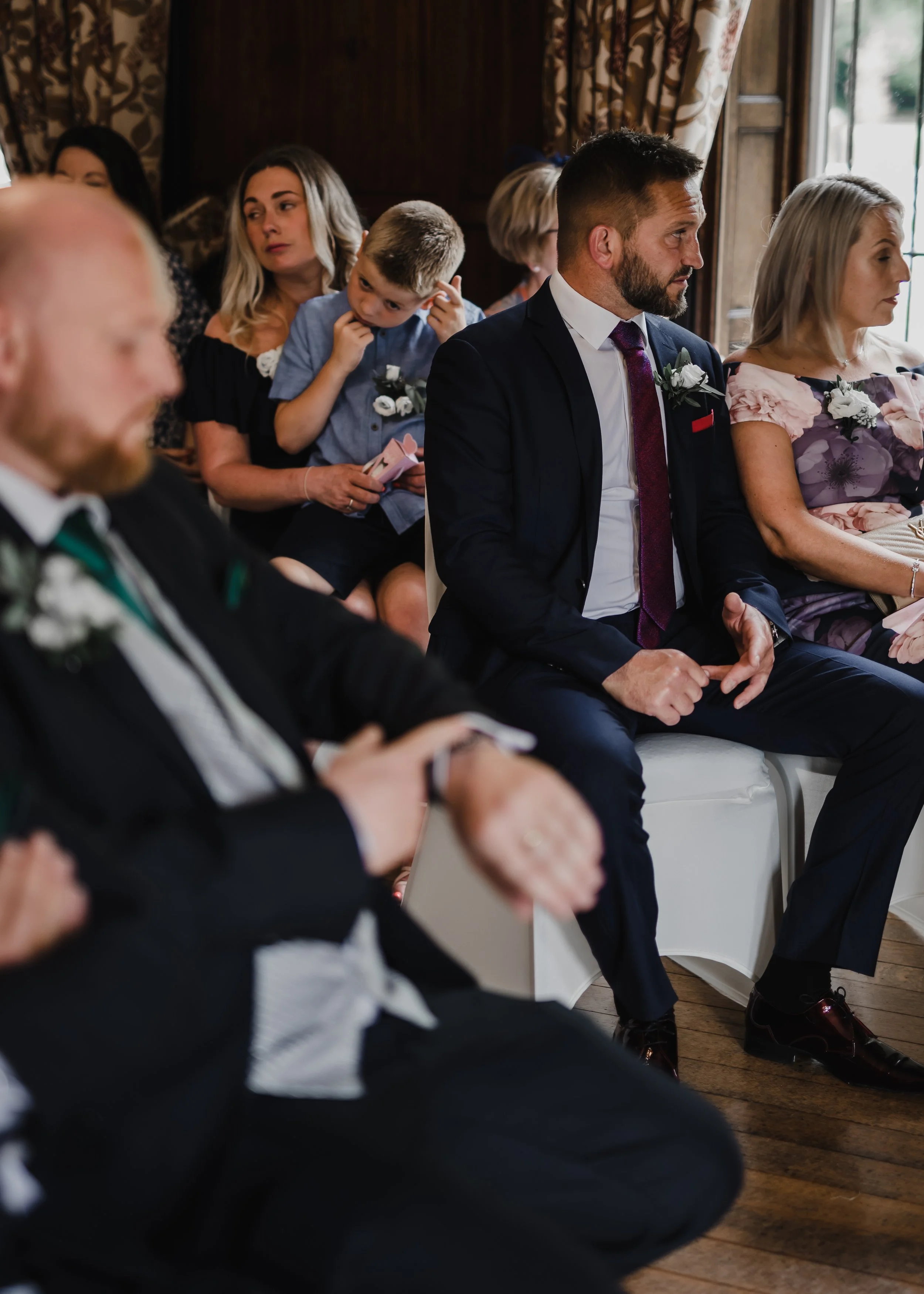 A group of people sitting at a formal event, possibly a wedding or ceremony, with some wearing suits with boutonnieres. The scene is indoors with wooden floors and floral curtains.