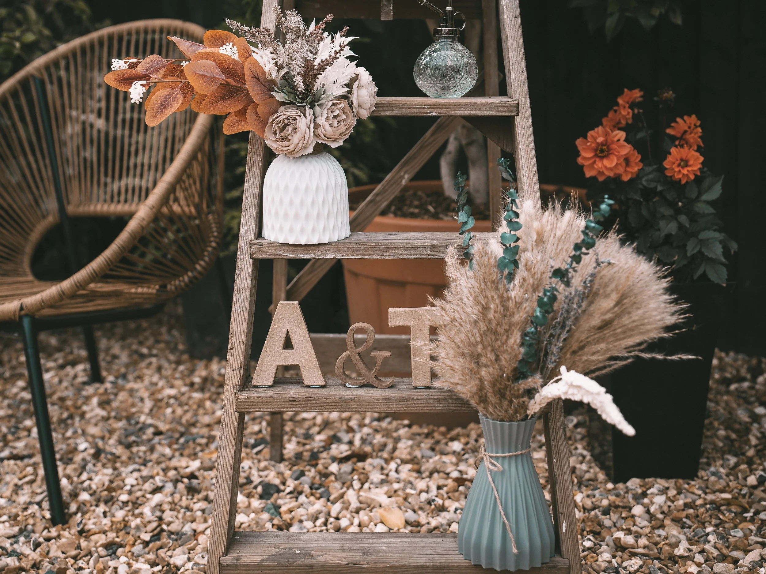 A rustic outdoor display featuring a wooden ladder with a white textured vase holding a bouquet of flowers and foliage, a glass spray bottle, an arrangement of orange flowers, and a large blue vase with pampas grass, with a wicker chair and gravel gr