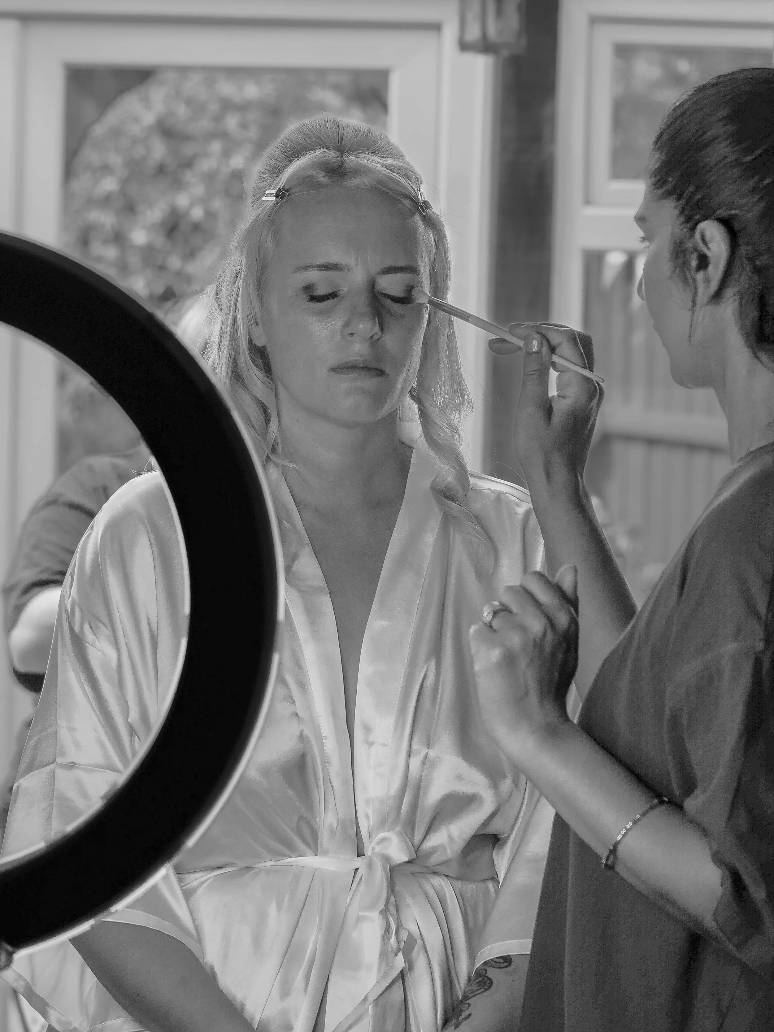 A woman in a medical or spa setting gets her makeup or eyelids done by another woman using a brush, with a mirror reflecting her face and a ring light partially visible in the foreground.