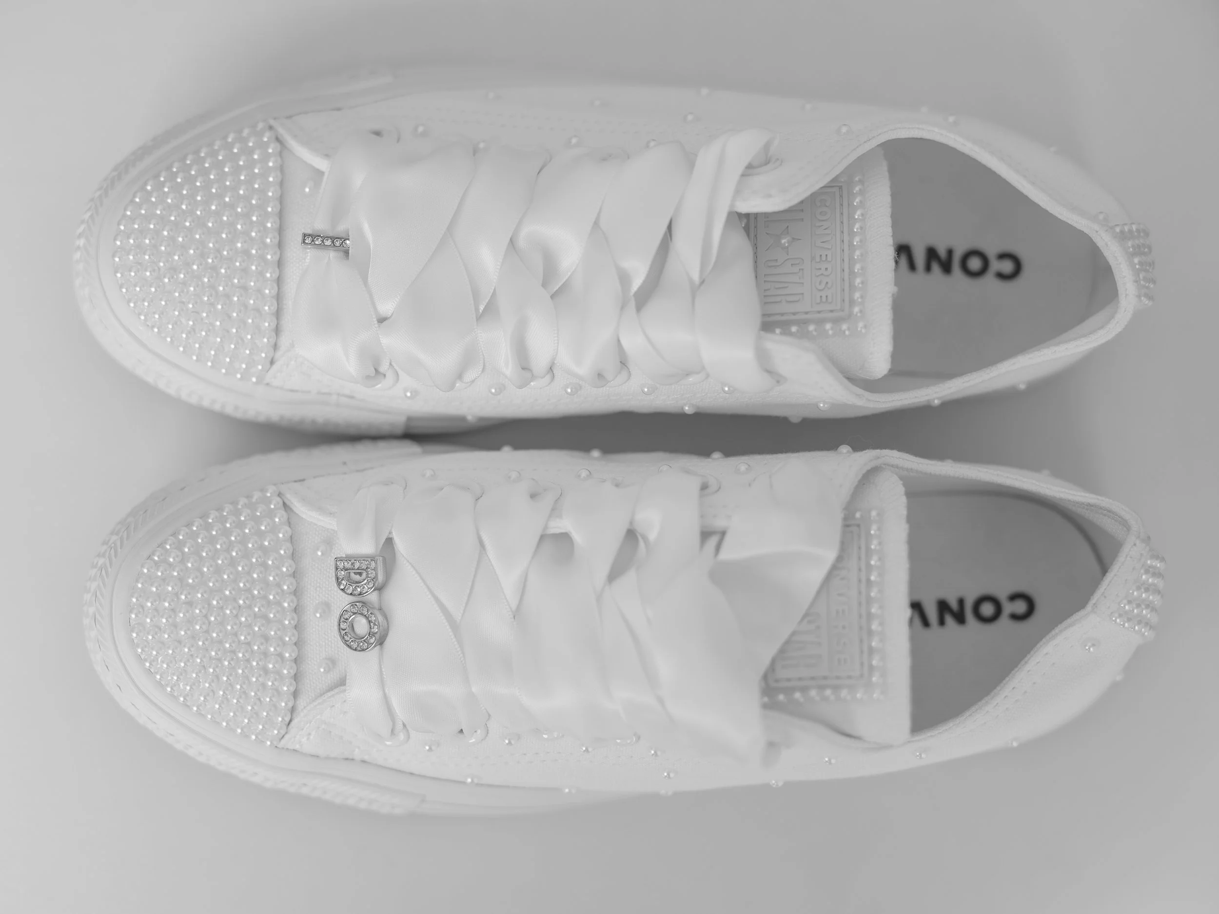 A pair of white sneakers decorated with small pearls and rhinestones, featuring satin ribbons for laces. The tongue of each sneaker shows the Converse logo.
