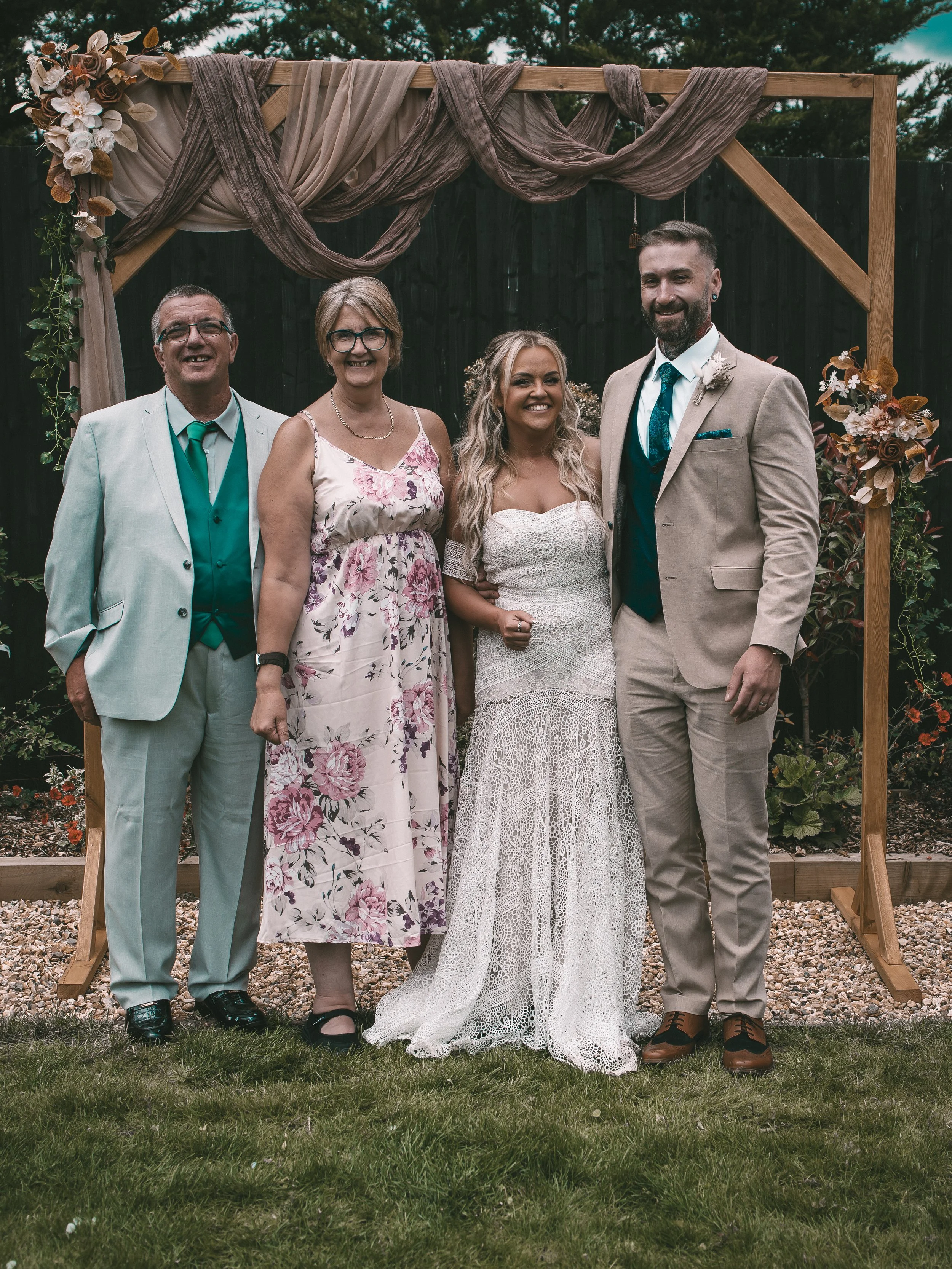 Five people standing outdoors in front of a wooden wedding arch decorated with fabric and flowers, dressed in formal attire, smiling at the camera.