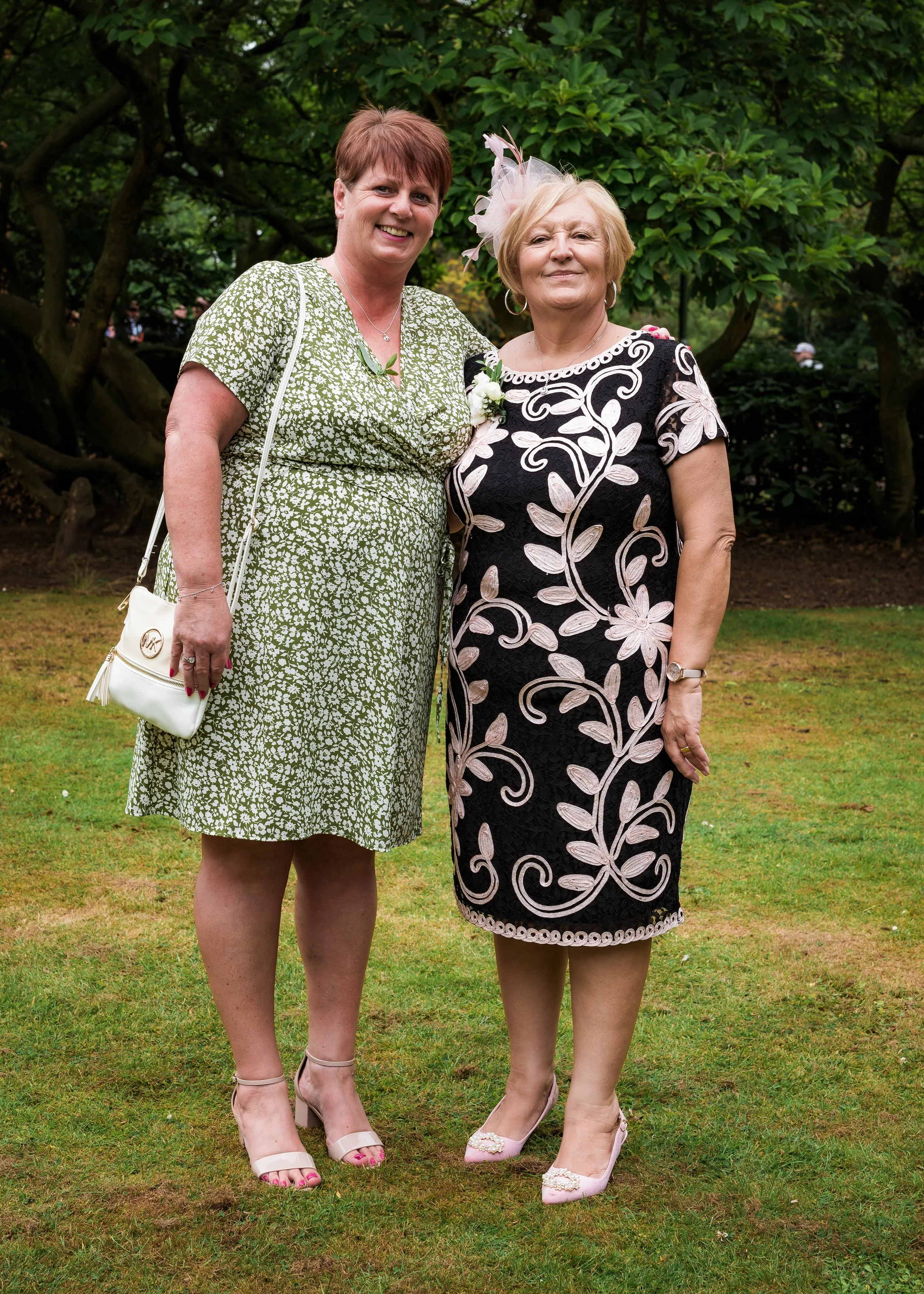 Two women standing outdoors on grass, smiling. One is wearing a green patterned dress with a white purse, the other is in a black dress with pink floral embroidery and a pink fascinator, both in heels.