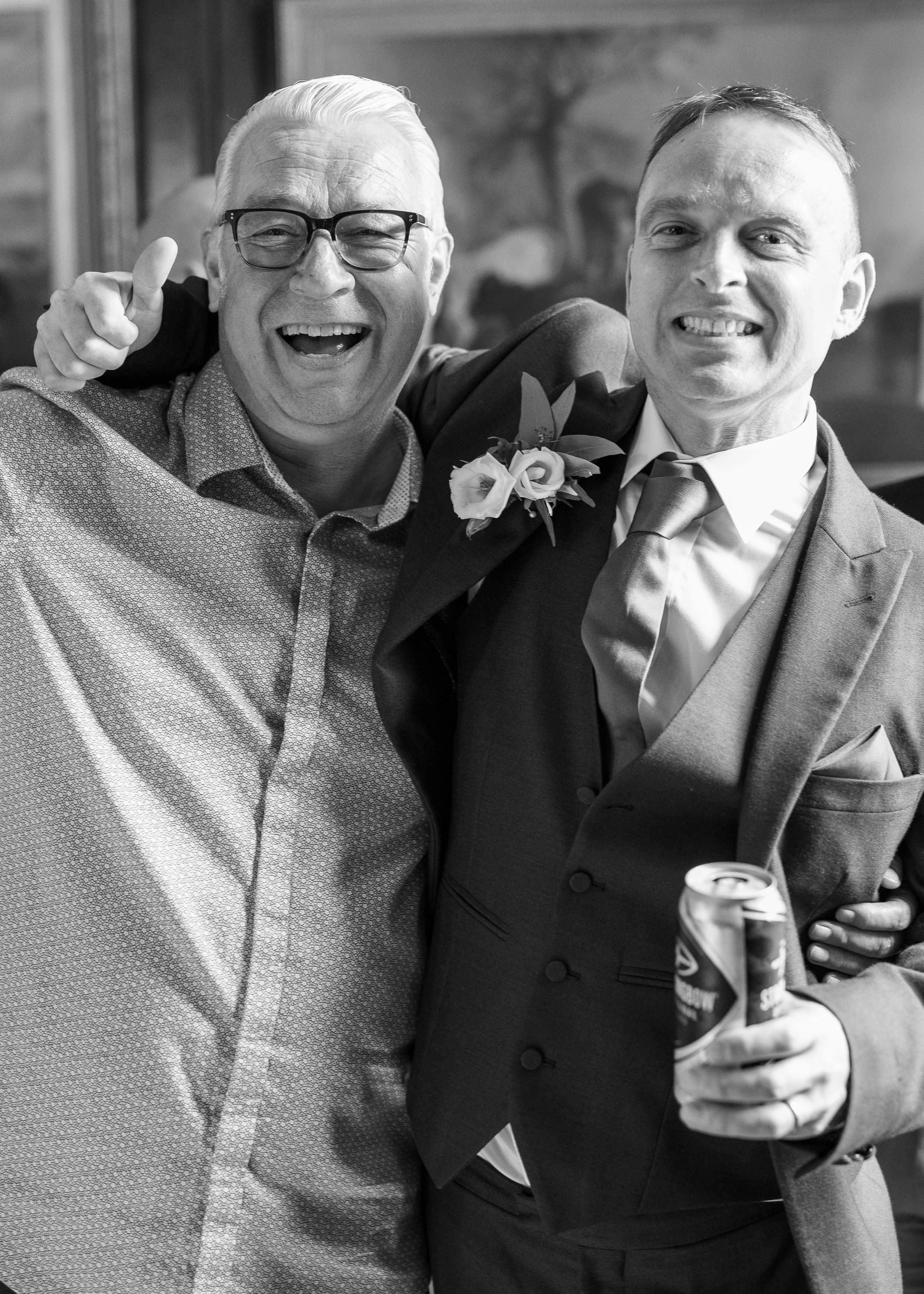 Two men at a celebration, one older with glasses and the other younger, both smiling and holding drinks, with an arm around each other.