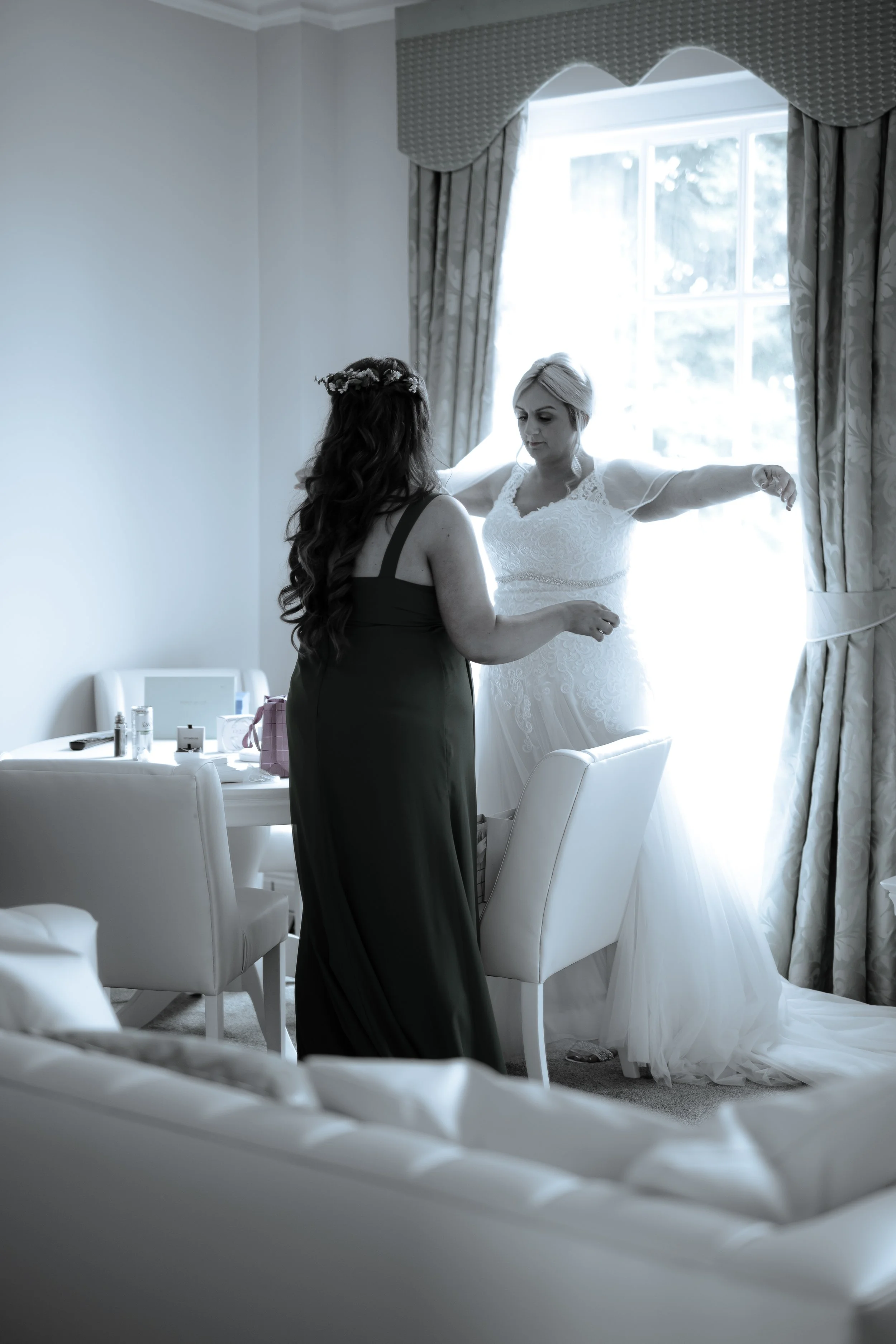 A bride in a wedding dress is being helped into her gown by another woman in a dark dress. They are in a bright room with a window and floral curtains, and there are various items on a table nearby.