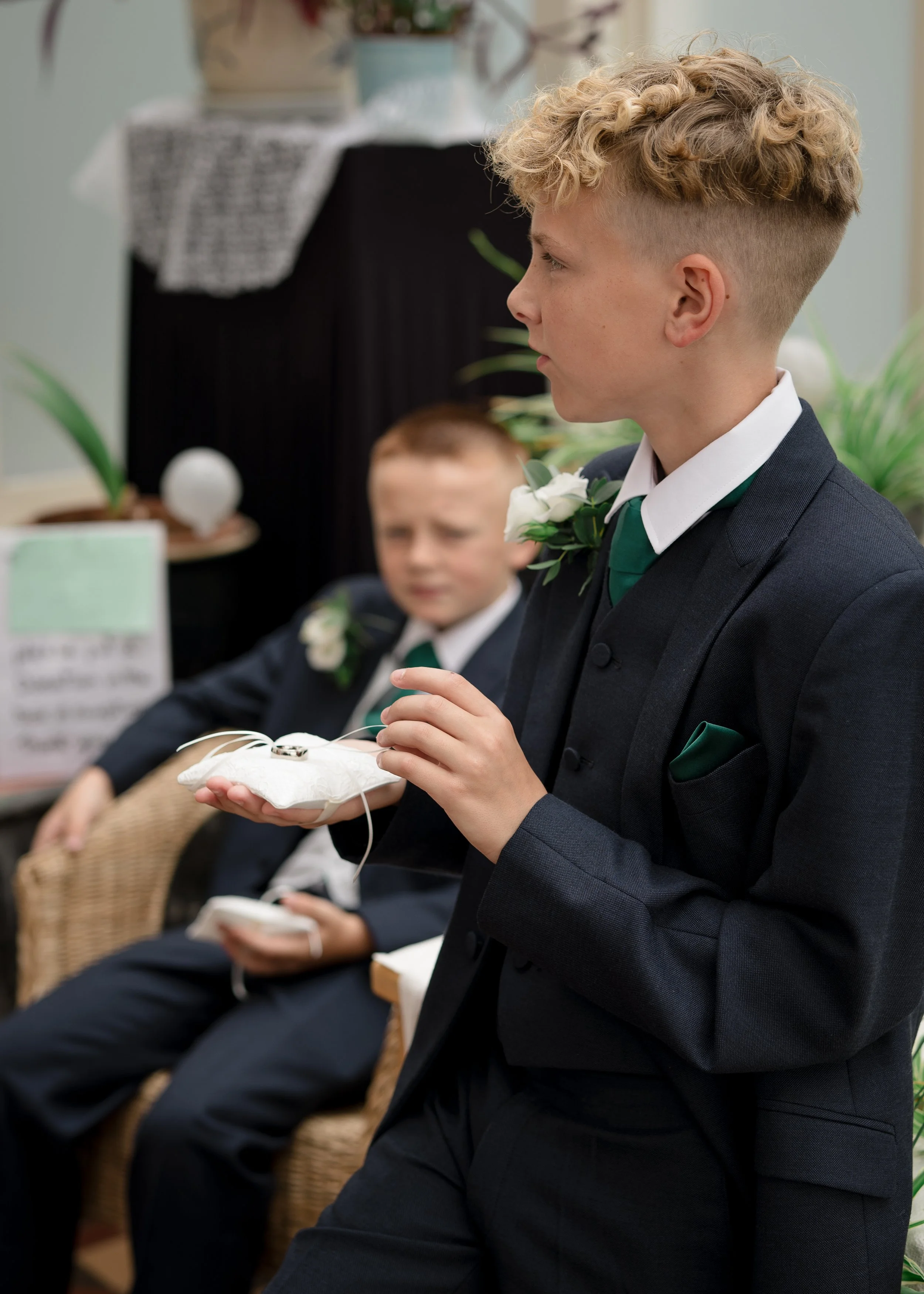 Young man in a formal suit participating in a wedding ceremony, holding a ring on a white cushion, with another young man seated beside him.