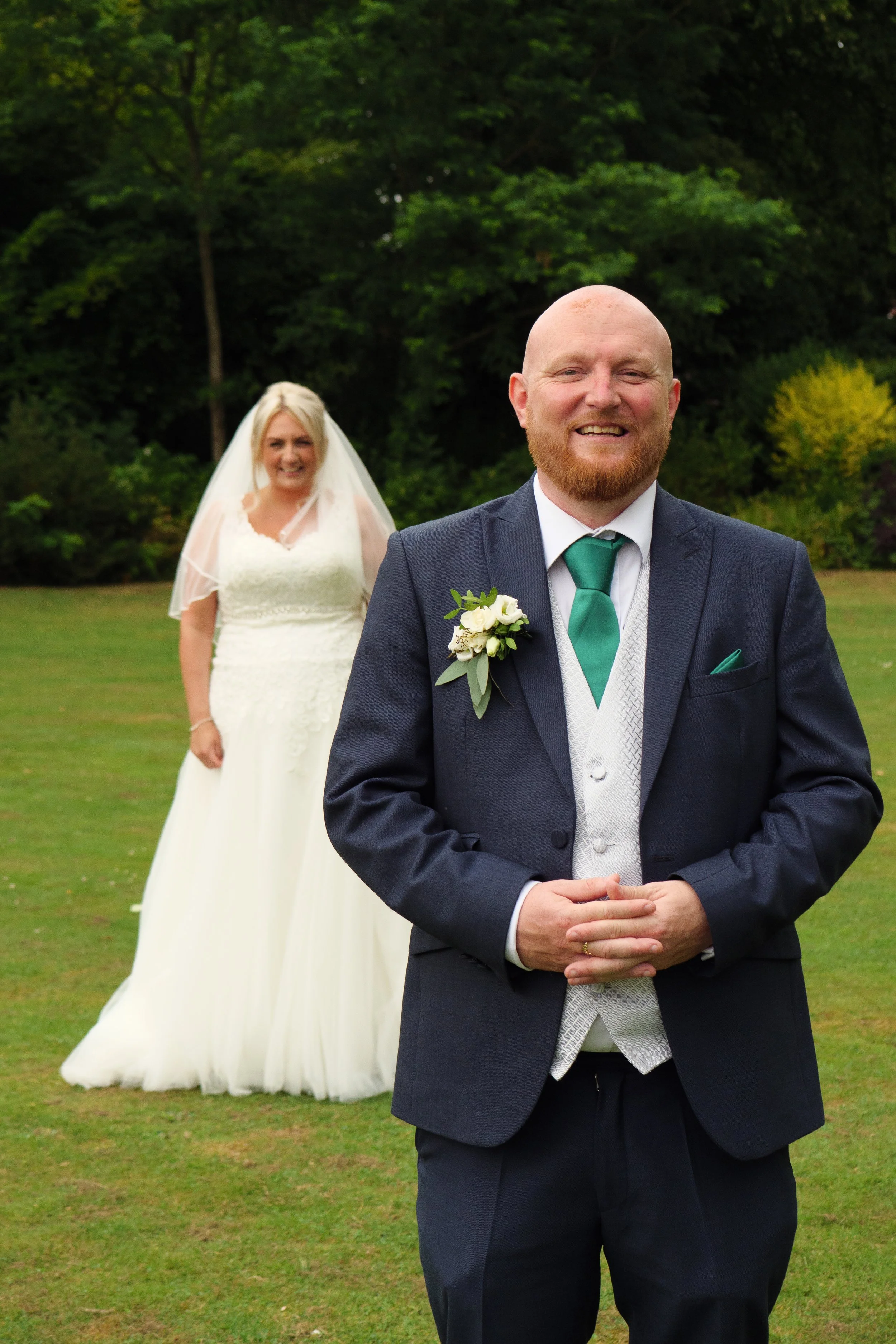 A bride in a white wedding dress and veil standing in the background, smiling, with a groom in a dark suit, white shirt, green tie, and boutonniere in the foreground, outdoors on a grassy area with trees.