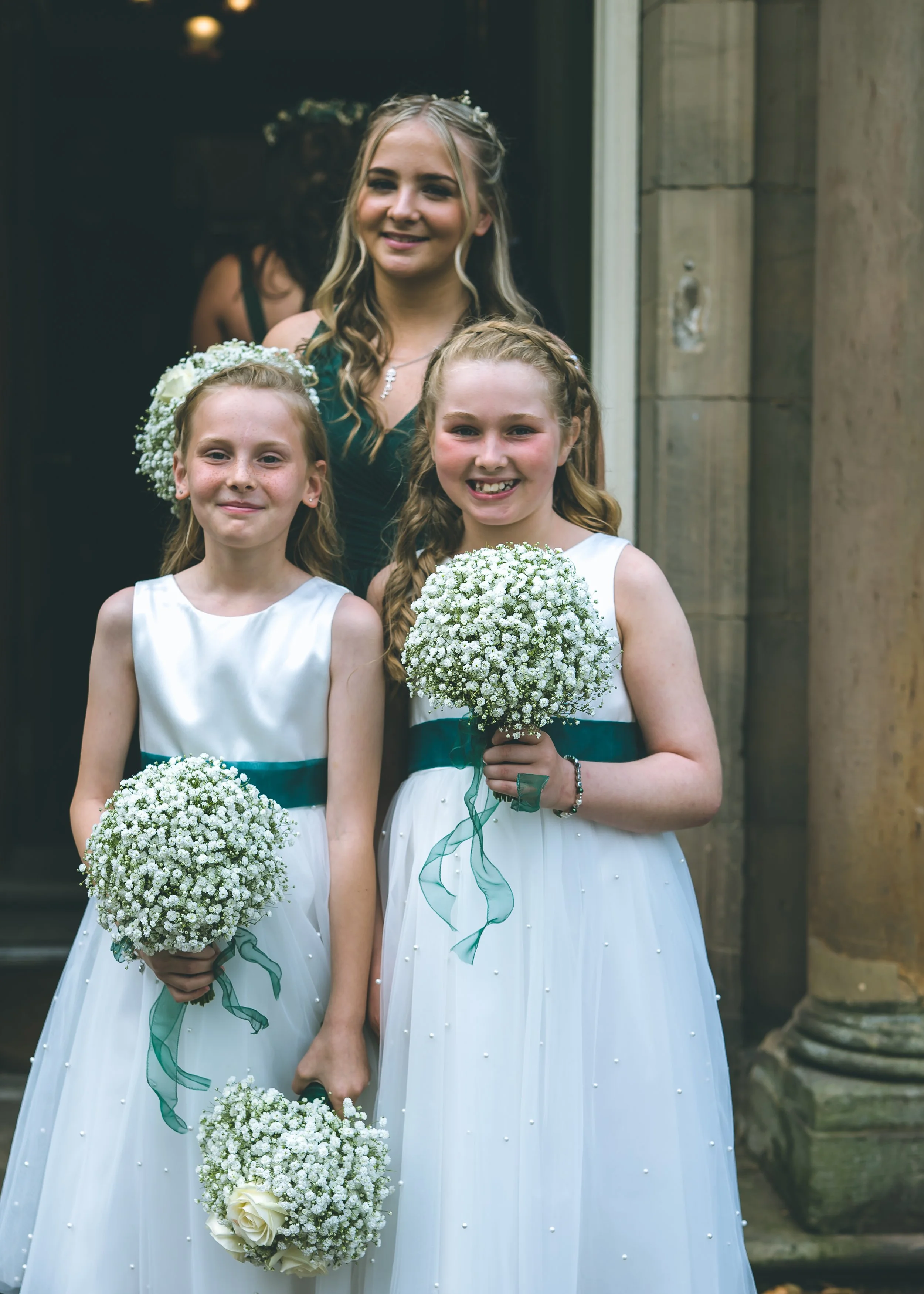 Three girls in white dresses holding small bouquets of white flowers, standing outside a building with stone columns. The girl in the center is smiling and has braided hair, the girl on the left is also smiling and wearing a floral headband, and the 