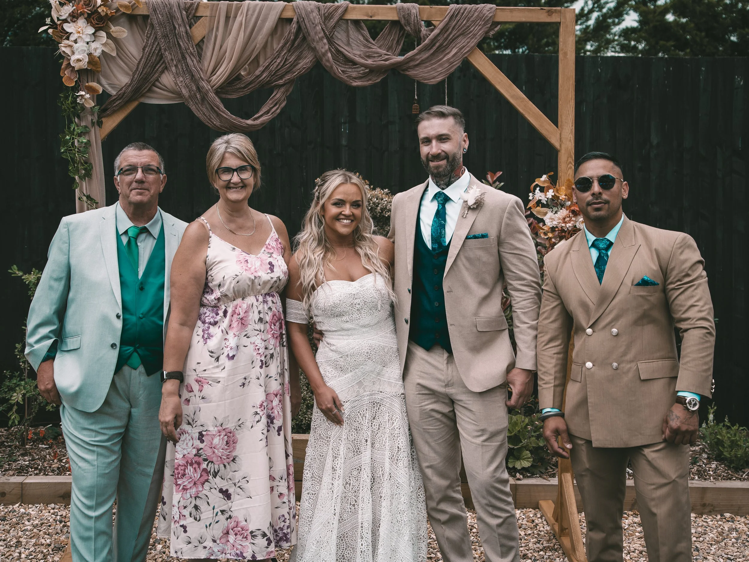 A group of six people standing outdoors in front of a wooden wedding arch adorned with fabric and flowers. The group includes four men and two women, all dressed in formal attire, smiling at the camera.