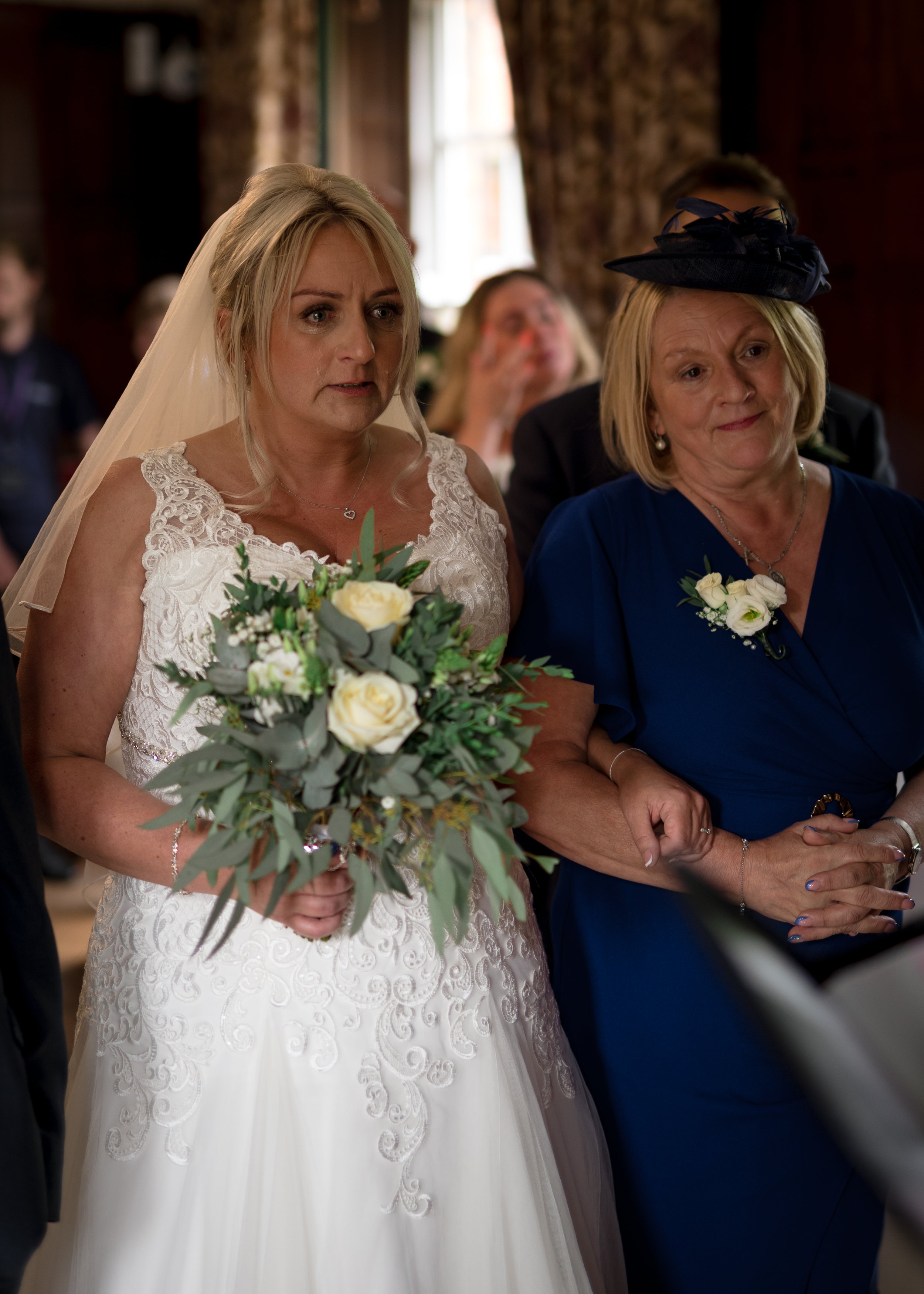 A bride in a white wedding dress holding a bouquet of white roses and greenery, standing next to an older woman in a blue dress and matching hat, both attending a wedding ceremony with other guests in the background.