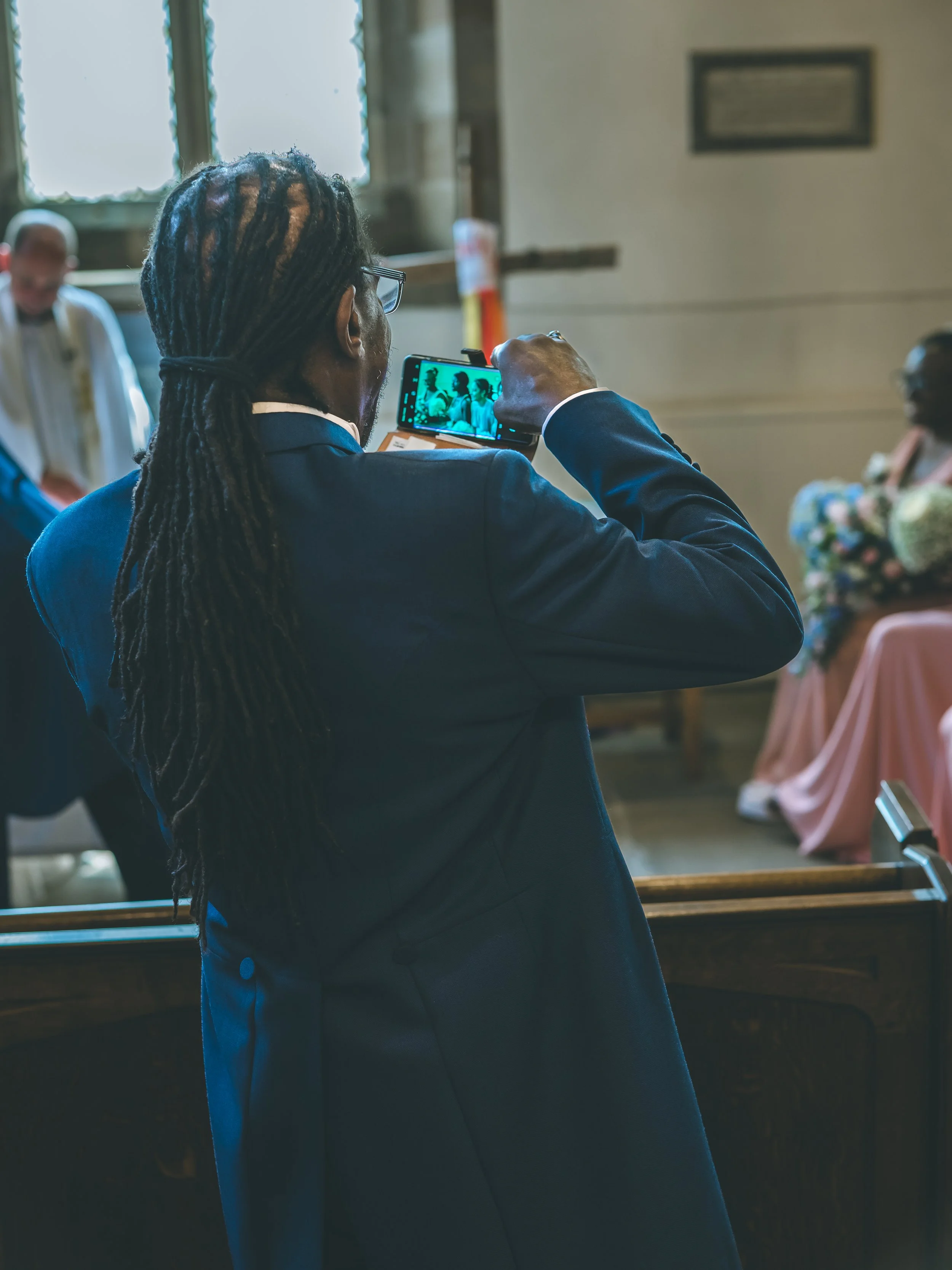 A person with long dreadlocks and glasses takes a photo of a group of people inside a church with a smartphone. The church interior has large windows with natural light, and the group is seated at tables with pink tablecloths.