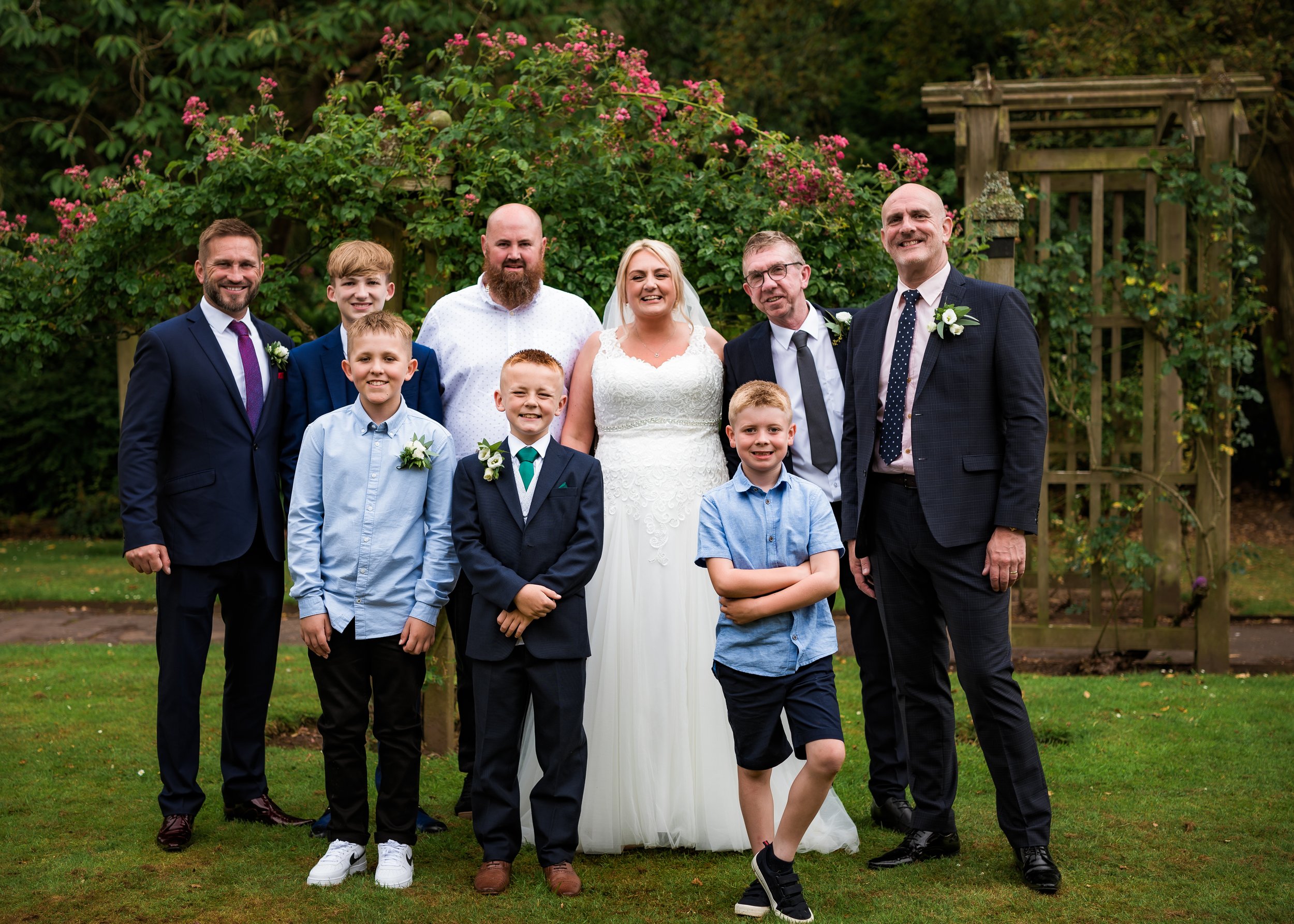 A group of ten people, including adults and children, dressed in formal and semi-formal attire, standing outdoors in front of pink flowering shrubs and wooden structures, celebrating a wedding.