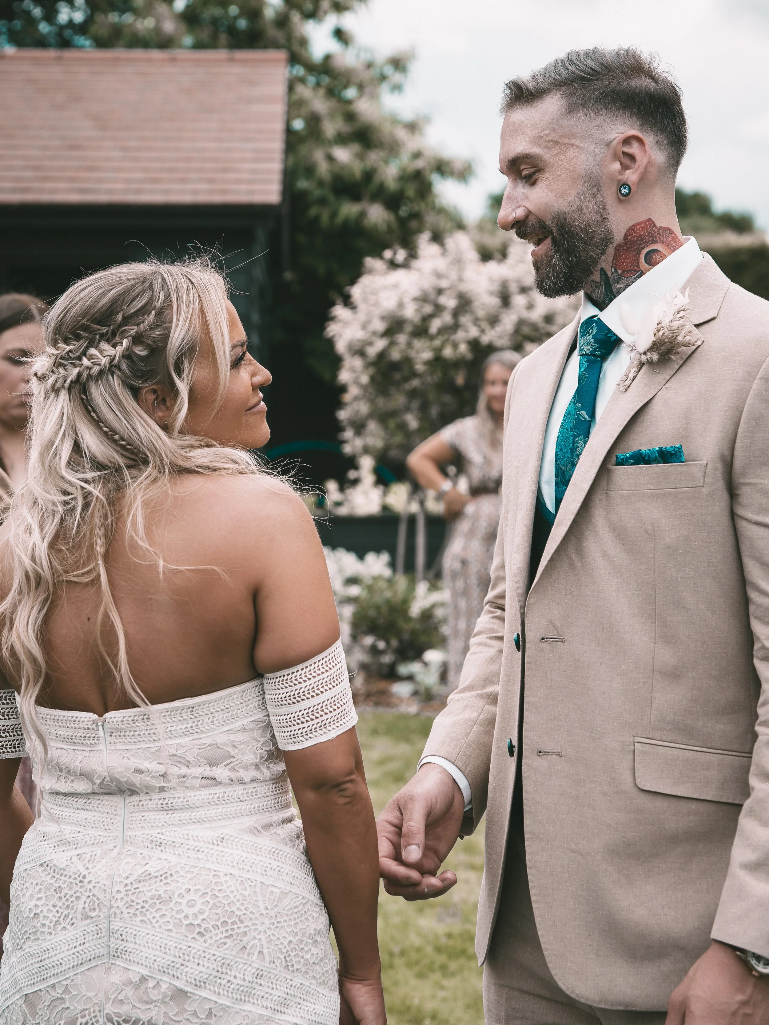 A bride and groom holding hands during their wedding ceremony, standing outdoors with guests in the background.
