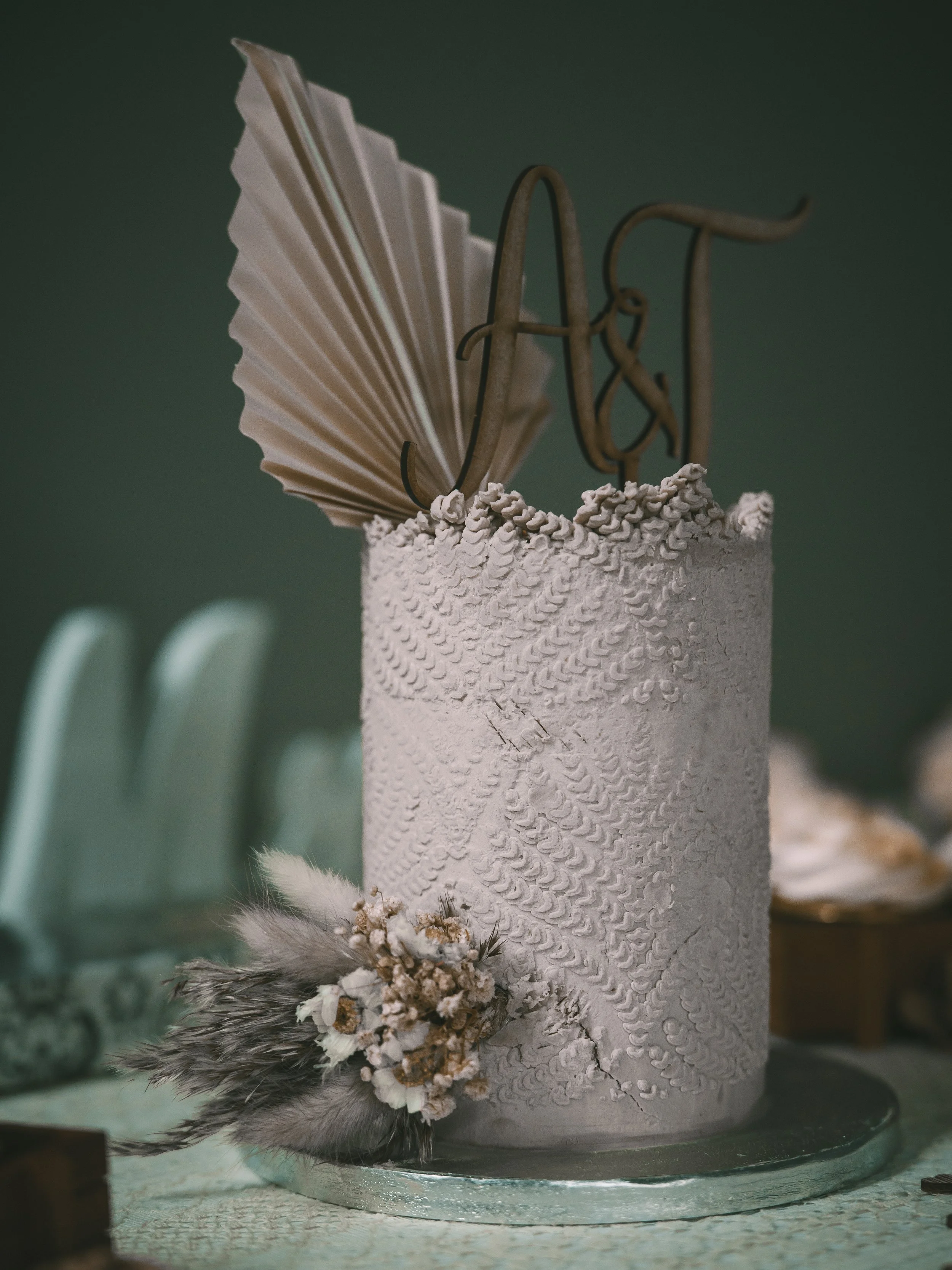 A cake decorated with white textured icing, dried flowers, feathers, and a large metallic 'A' topper, with a folded paper fan and a small arrangement of dried flowers at the base.