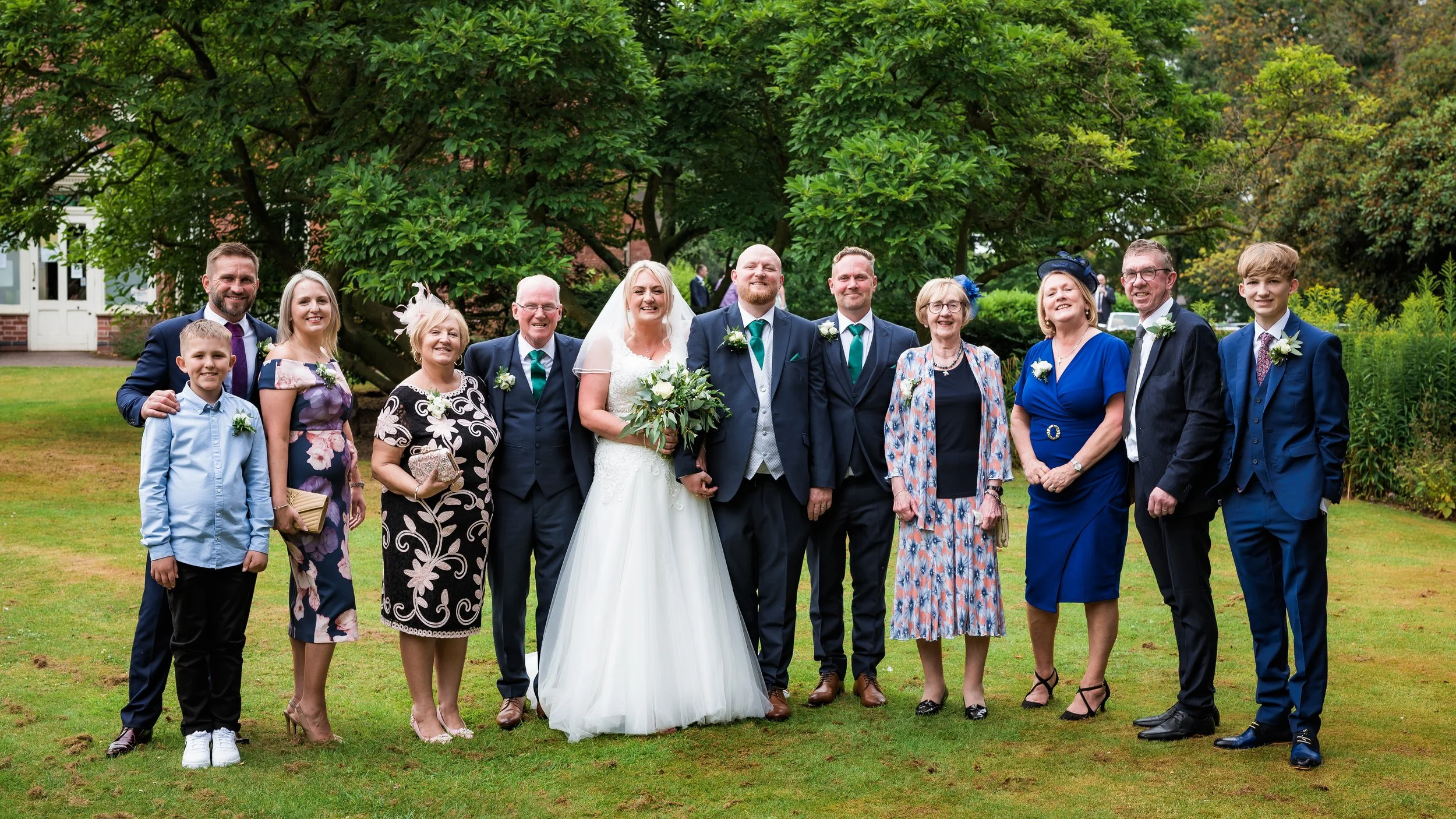 A large group of people, including a bride in a white wedding gown and others in formal attire, posing for a photo outdoors in a garden with green trees. The group appears to be celebrating a wedding.